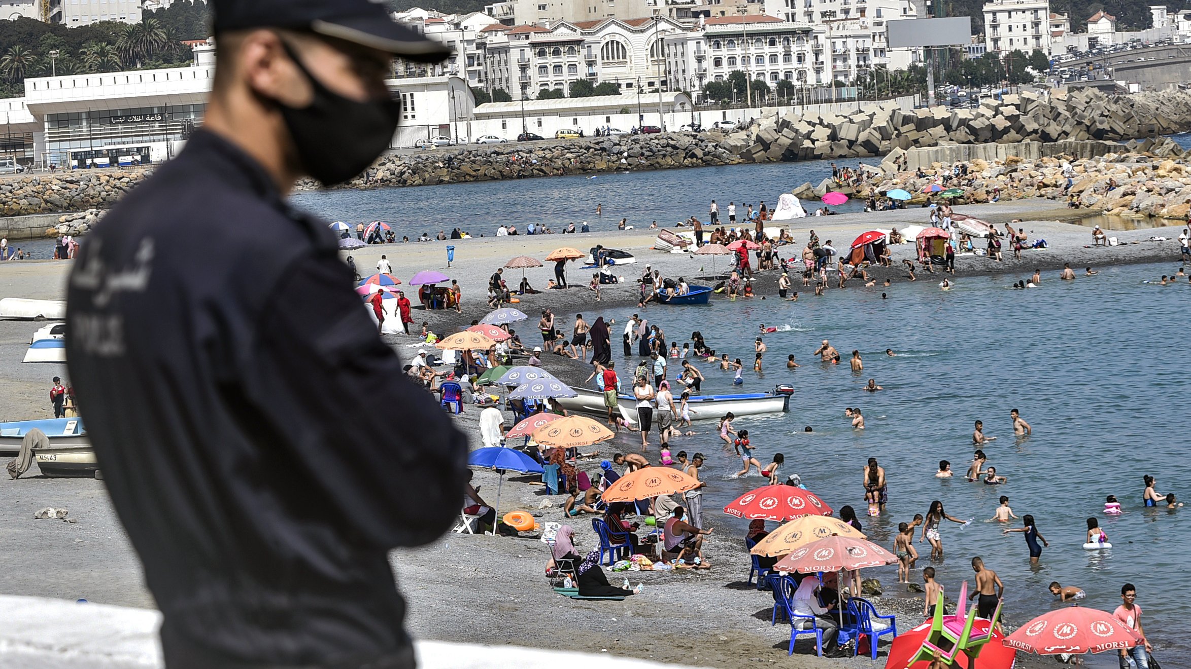 L'image montre un agent de sécurité en premier plan, portant un masque, surveillant une plage animée. En arrière-plan, des familles et des groupes de personnes profitent du temps ensoleillé, se baignant dans l'eau et se reposant sous des parasols colorés. On aperçoit également des bateaux au loin et des bâtiments sur la côte qui ajoutent à l'ambiance estivale de la scène.