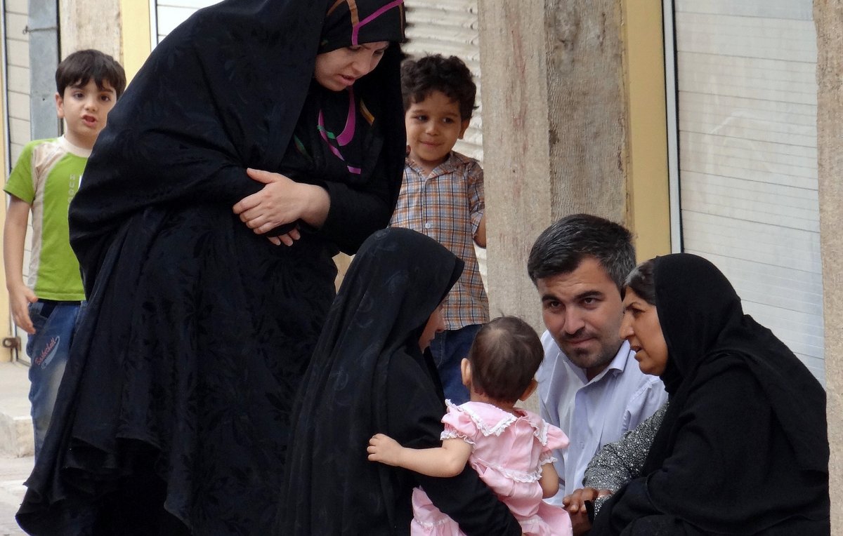 L'image montre un groupe de personnes dans une rue. Deux femmes, vêtues de vêtements noirs, sont assises sur le sol et discutent. L'une d'elles tient un bébé, qui porte une légère robe rose. À proximité, un homme regarde les femmes et semble participer à la conversation. Un jeune garçon, en arrière-plan, observe la scène. L'environnement apparaît urbain, avec des bâtiments en briques visibles. L'atmosphère semble conviviale et familiale.