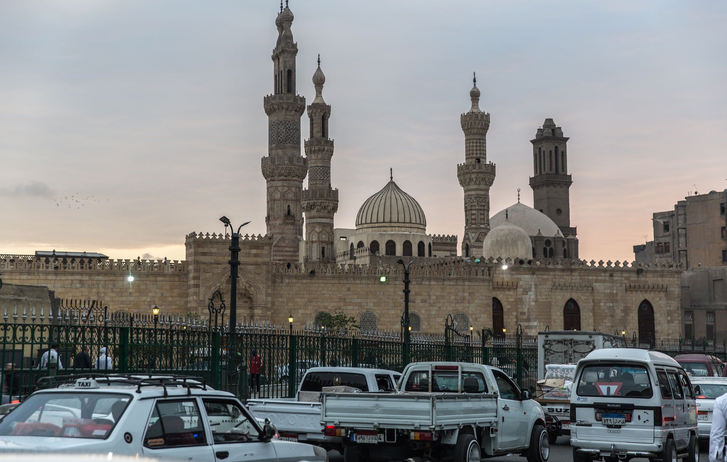 L'image montre un paysage urbain avec des bâtiments emblématiques, comprenant des mosquées avec des minarets élancés et des dômes caractéristiques. Au premier plan, on peut voir des véhicules traversant une route animée, entourés d'une clôture verte. L'éclairage doux de la fin de journée ajoute une atmosphère chaleureuse à la scène, tandis que le ciel présente des teintes nuancées, suggérant le coucher du soleil. L'architecture évoque un style traditionnel, typique de certaines grandes villes historiques.