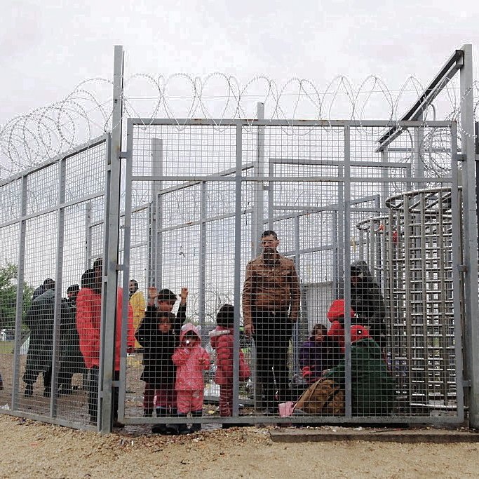 L'image montre un groupe de personnes à l'intérieur d'une structure grillagée, entourée de barbelés. On peut voir des adultes et des enfants, certains portant des vêtements colorés, semblant attendre dans un espace très restreint. L'ambiance semble préoccupante, suggérant une situation de confinement ou d'attente difficile. Le ciel est nuageux, ce qui contribue à une impression de morosité.