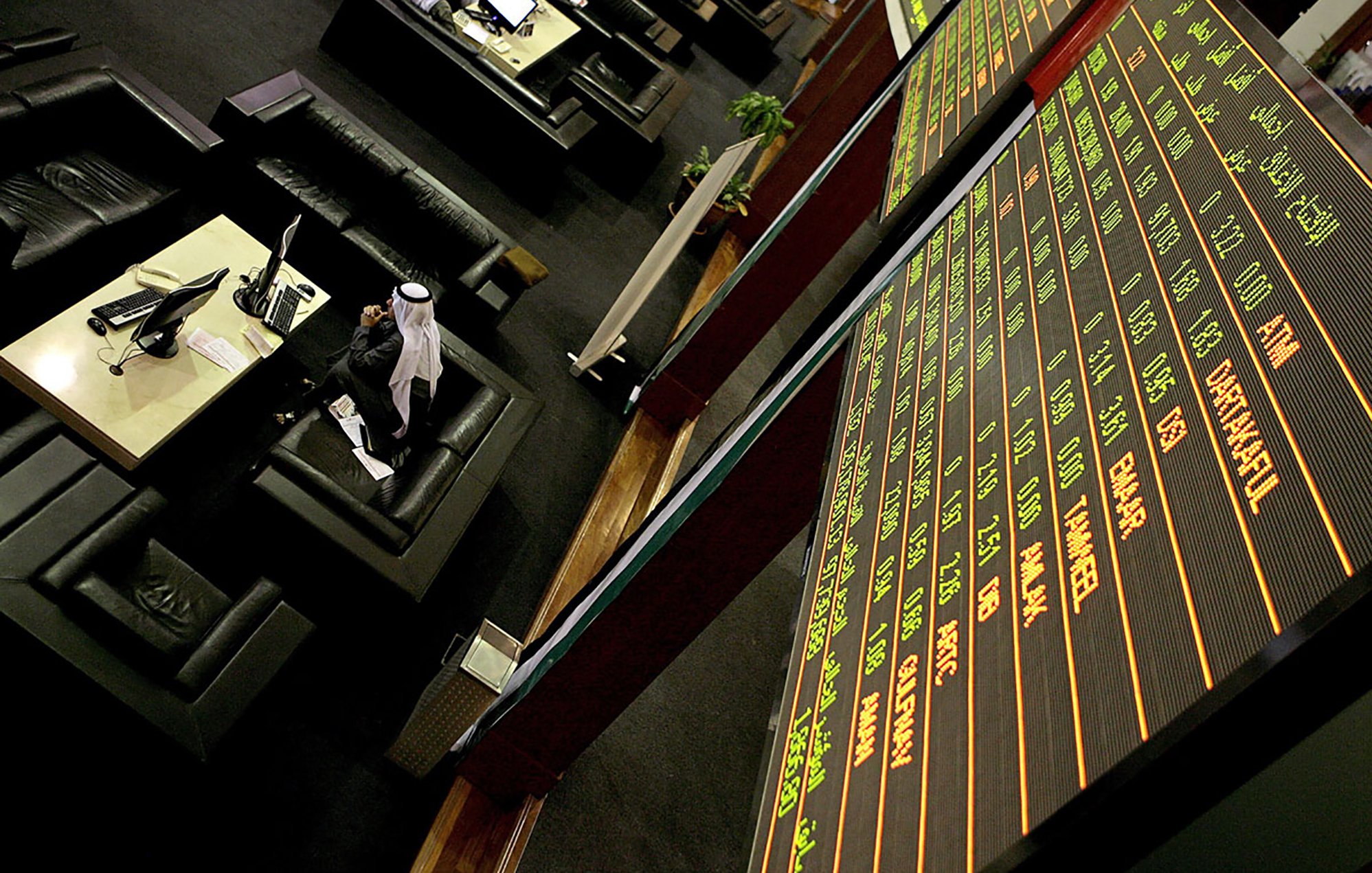 L'image montre un environnement de bourse avec des tables et des sièges confortables. À l'arrière-plan, un grand tableau électronique affiche des informations financières, comme des chiffres et des noms de sociétés. On peut voir quelques personnes, dont un homme en tenue traditionnelle, probablement en train de suivre les fluctuations du marché. L'ambiance semble professionnelle et concentrée, caractéristique d'une salle de négociation.