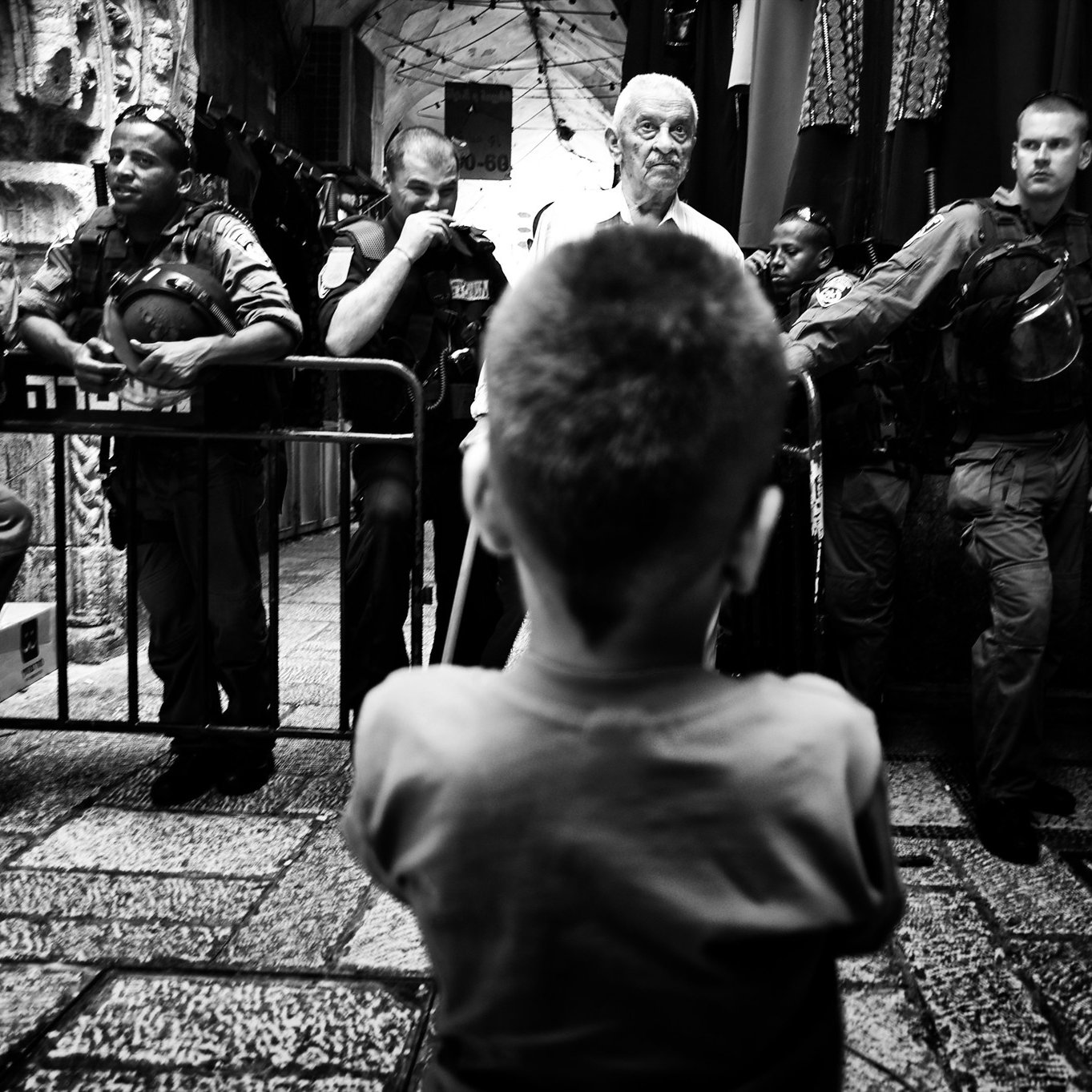 این تصویر سیاه و سفید، یک صحنه یاری را نشان می‌دهد که در آن یک کودک در حالت تماشا، به دو مرد بزرگ‌سال و یک گروه از نیروهای امنیتی نگاه می‌کند. کودک از پشت تصویر دیده می‌شود و توجه او به مردی که در مقابلش ایستاده جلب شده است. نیروهای امنیتی در کنار آنها قرار دارند و به نظر می‌رسد که صحنه‌ای از تنش یا تعامل میان افراد در حال رخ دادن است. فضای تصویر حس تنش و توجه را به خوبی منتقل می‌کند.