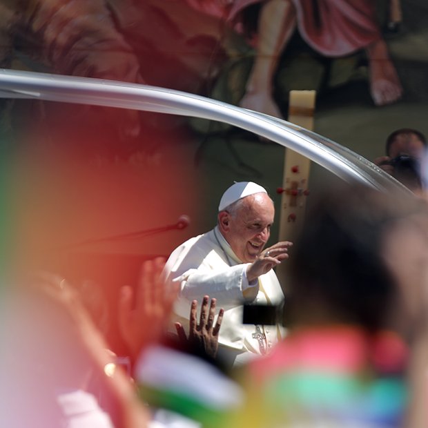 L'image montre une scène d'un événement public avec le Pape François. Il est souriant et fait un geste de la main en saluant la foule. On peut apercevoir des drapeaux multicolores dans le premier plan, ce qui indique une ambiance festive et dynamique. En arrière-plan, on distingue des illustrations ou des représentations qui semblent évoquer une célébration ou un rassemblement spirituel. L'ensemble dégage une atmosphère de joie et d'enthousiasme de la part des personnes présentes.