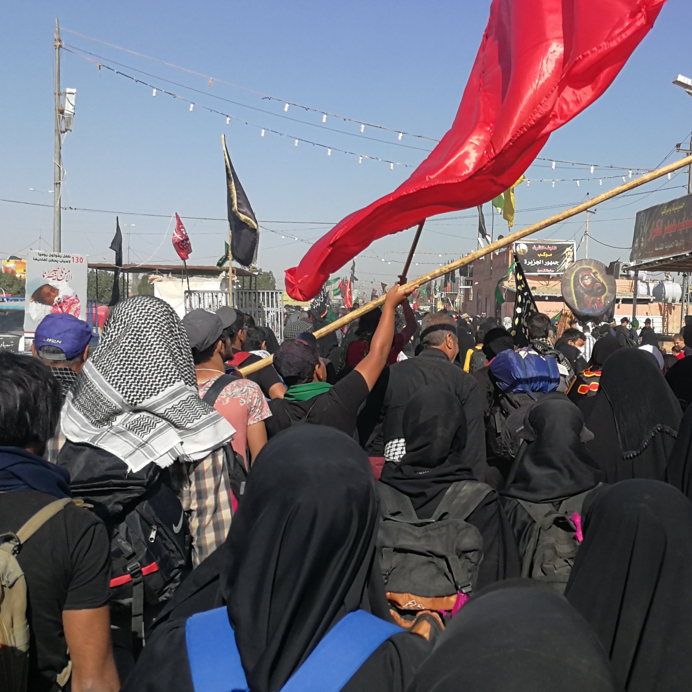 تظهر في الصورة مجموعة كبيرة من الأشخاص، معظمهم يرتدون الملابس السوداء، مما يشير إلى أنهم ربما يشاركون في مناسبة دينية. يحمل بعضهم أعلامًا حمراء، وهناك لافتات وأعلام أخرى في الخلفية. الأجواء تظهر حماسًا وجمعًا كبيرًا من الناس، مما يعكس أهمية الحدث الذي يشتركون فيه. السماء صافية وأشعة الشمس تضيء المكان.