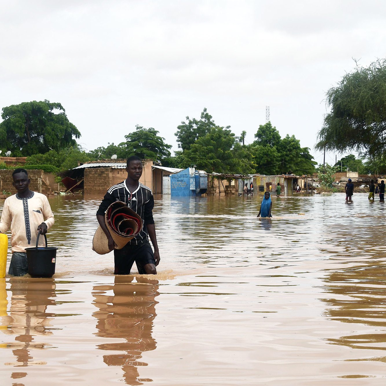 تظهر الصورة مشهداً لمنطقة تعرضت للفيضانات، حيث يغمر الماء الأرض بشكل كبير. يوجد شخصان يسيران في المياه العميقة، يحمل أحدهما دلوًا ويبدو أنهما يجمعان بعض الممتلكات. في الخلفية، يمكن رؤية أكواخ وأشجار، مما يدل على أن المنطقة قد تضررت. السماء ملبدة بالغيوم، مما يشير إلى ظروف جوية غير مستقرة. يعكس المشهد الصعوبات التي يواجهها السكان نتيجة الفيضانات.