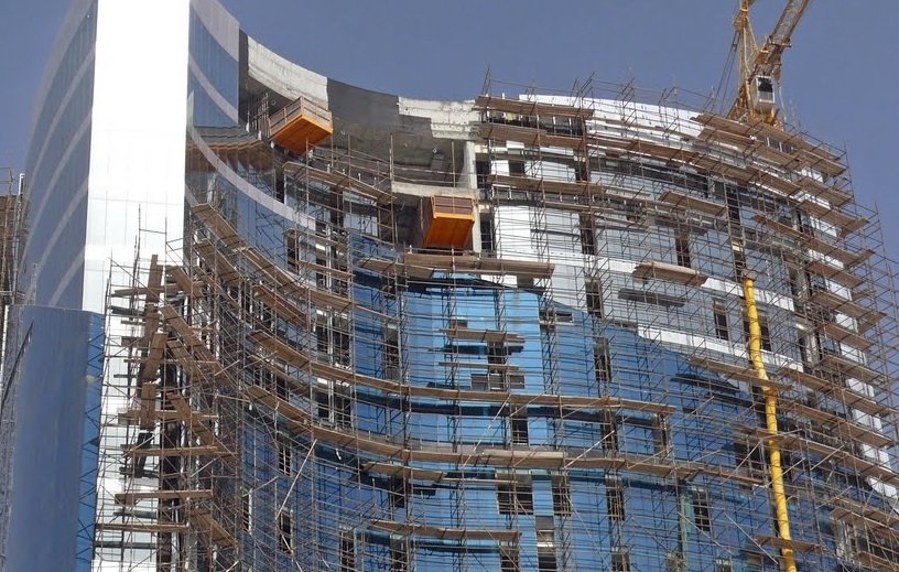 L'image montre un bâtiment en construction. On peut voir une structure moderne avec des façades en verre, entourée d'un échafaudage en métal. Plusieurs niveaux sont visibles, et des éléments de construction, comme des grues, sont présents pour faciliter les travaux. Le ciel est dégagé, ce qui indique une belle journée.