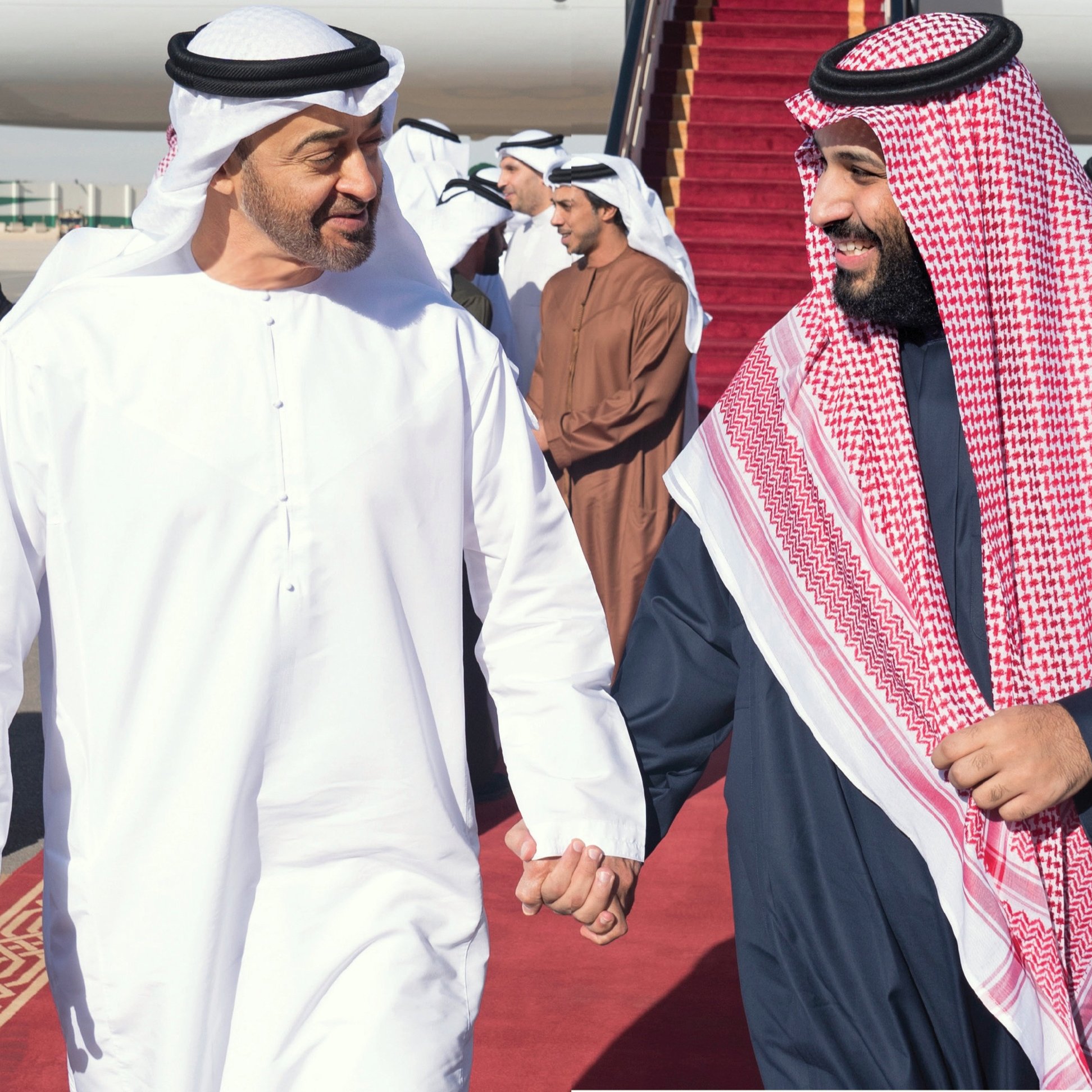 L'image montre deux hommes vêtus de vêtements traditionnels arabes, marchant côte à côte sur un tapis rouge. Ils semblent échanger un moment amical, souriant l'un à l'autre. En arrière-plan, plusieurs autres personnes sont présentes, ainsi qu'un avion stationné. L'atmosphère semble chaleureuse et détendue.