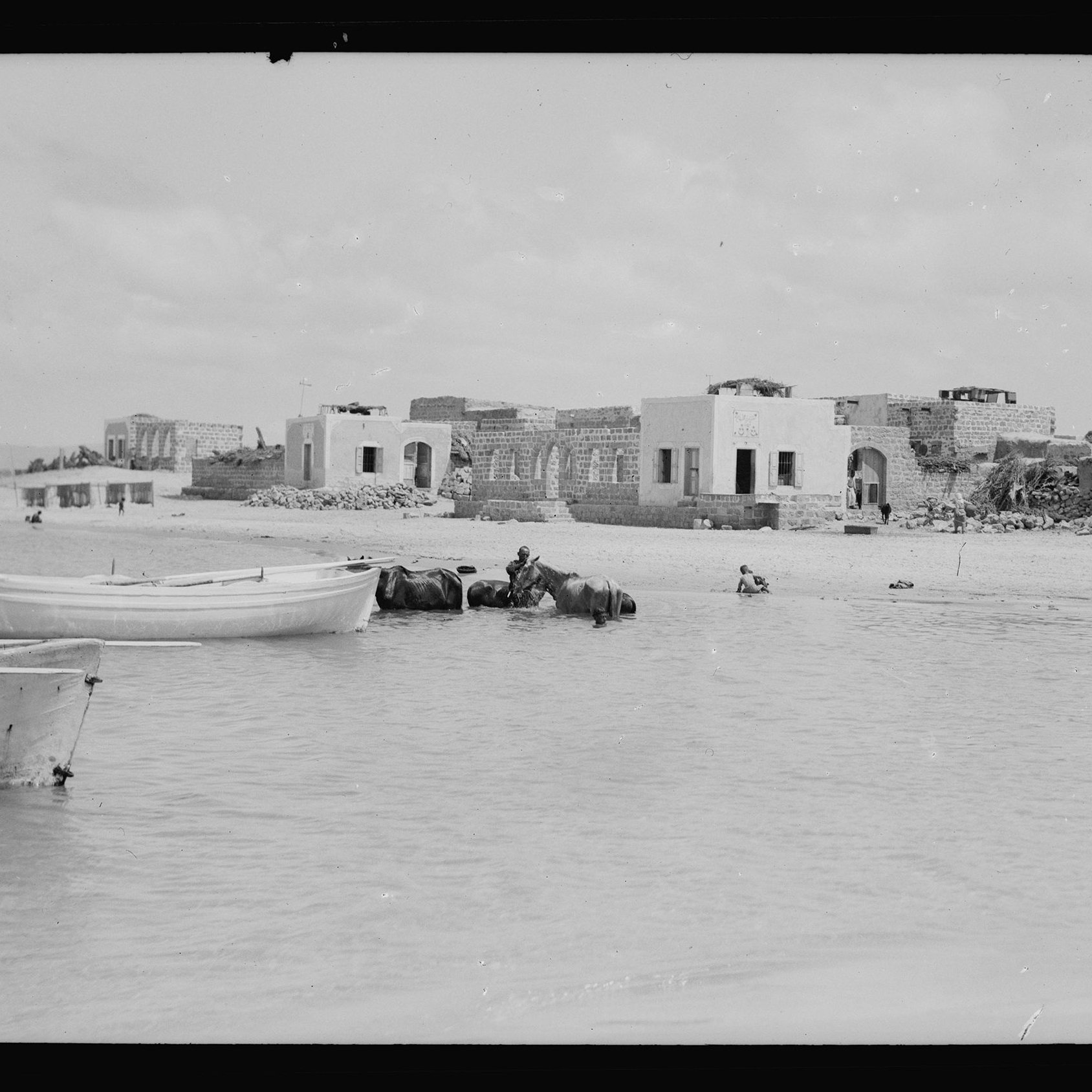 L'image montre une scène maritime où plusieurs bateaux, probablement des barques, sont amarrés au bord d'une eau peu profonde. En arrière-plan, on peut apercevoir des bâtiments en ruines, suggérant une ancienne architecture. À proximité, un homme semble se trouver dans l'eau, tandis qu'un autre est à cheval ou monté sur un animal aquatique. Le ciel est nuageux, apportant une lumière diffuse à l'ensemble de la scène. L'atmosphère semble tranquille et un peu nostalgique, évoquant un passé révolu.