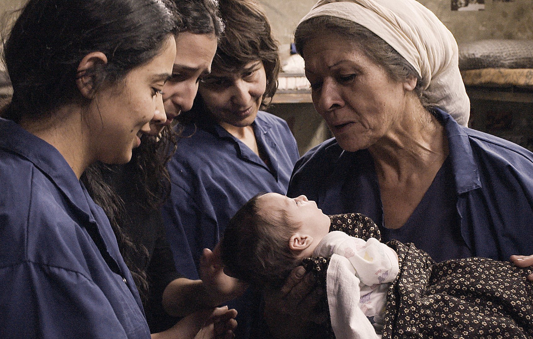 L'image montre un moment touchant où une femme âgée tient un bébé dans ses bras, partageant une interaction affectueuse. Autour d'elle, trois jeunes femmes observent et semblent émerveillées par ce moment. Elles portent toutes des vêtements similaires, de couleur bleue, et l'environnement semble simple et chaleureux. L'atmosphère dégage une sensation de tendresse et de solidarité entre les générations.