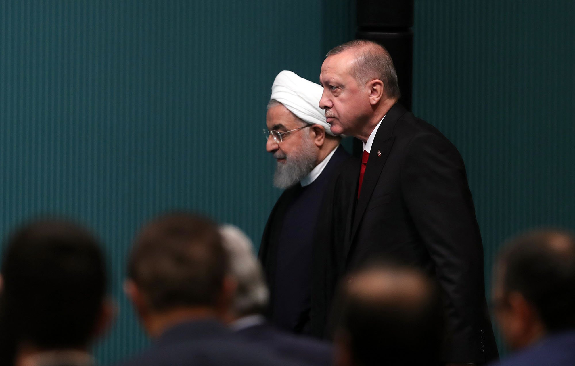 The image shows two men walking side by side. One is wearing a white turban and a black robe, while the other is dressed in a formal suit. They appear to be in a formal setting, likely a conference or meeting, with other individuals partially visible in the foreground. The background features a minimalist wall with a light blue-green color. The atmosphere seems serious and professional.