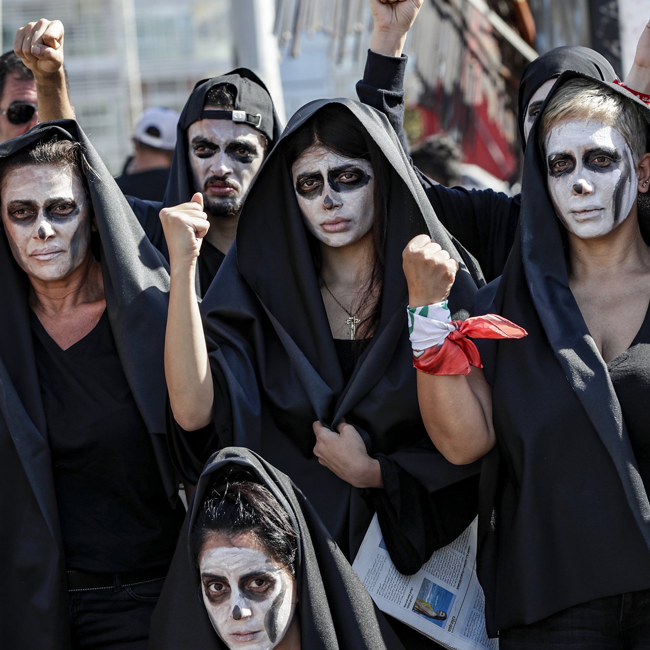 L'image montre un groupe de personnes, principalement des femmes, portant des vêtements noirs et des capes. Leur visage est peint en blanc avec des accents noirs, et elles levèrent les poings de manière déterminée. L'atmosphère semble être celle d'une manifestation ou d'une protestation, avec une ambiance de solidarité et de revendication. Certains participants portent un foulard ou un accessoire rouge. L'ensemble dégage un sentiment fort de résistance et d'engagement.