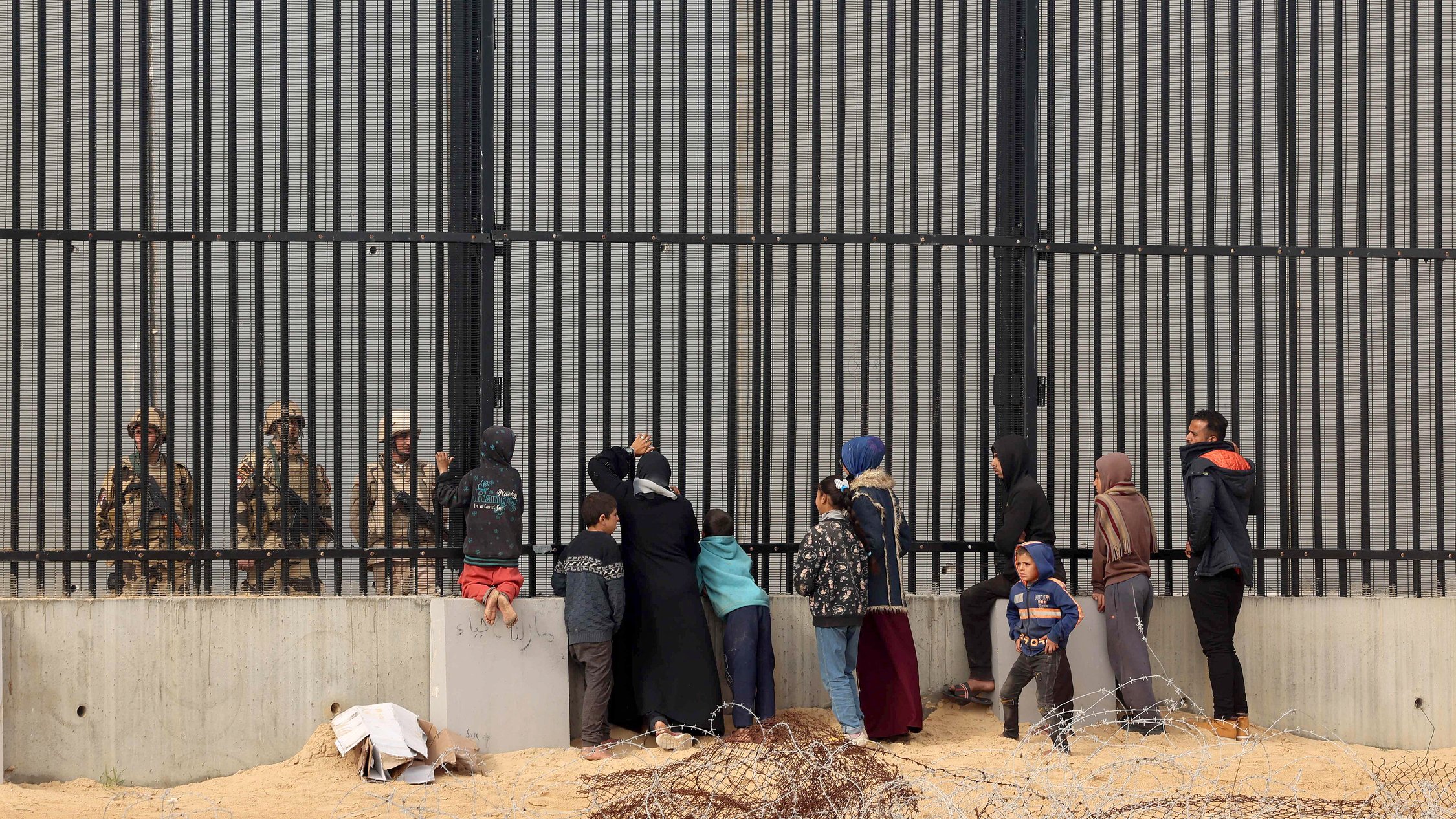 The image depicts a group of people, including women and children, standing in front of a tall, barred fence. On the other side of the fence are soldiers or guards, who appear to be watching the group. The scene conveys a sense of separation and tension, with barbed wire in the foreground emphasizing the barriers between the two sides. The individuals on the left seem to be engaging with the guards, possibly seeking assistance or communication. The overall atmosphere suggests a complex and challenging situation, likely related to migration or security issues.