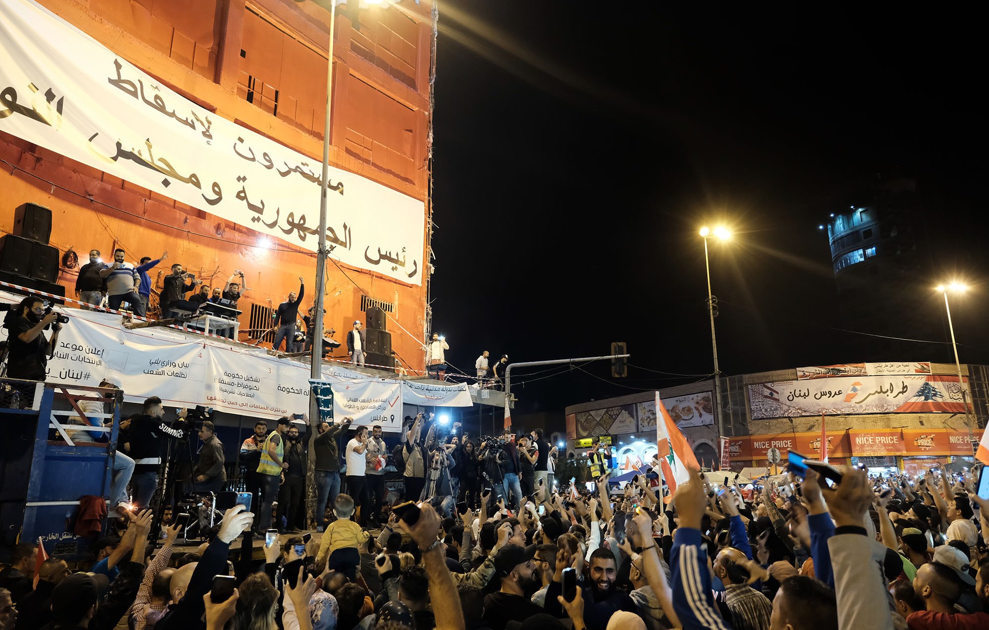 L'image montre une grande foule rassemblée dans une place publique, manifestant avec enthousiasme. Les gens lèvent les mains et brandissent des drapeaux, exprimant des émotions fortes. À l'arrière-plan, on peut voir un grand panneau affichant des messages politiques. Des personnages sont visibles sur une scène surélevée, semblant s'adresser à la foule. L'ambiance semble festive et dynamique, avec des lumières qui illuminent la scène.