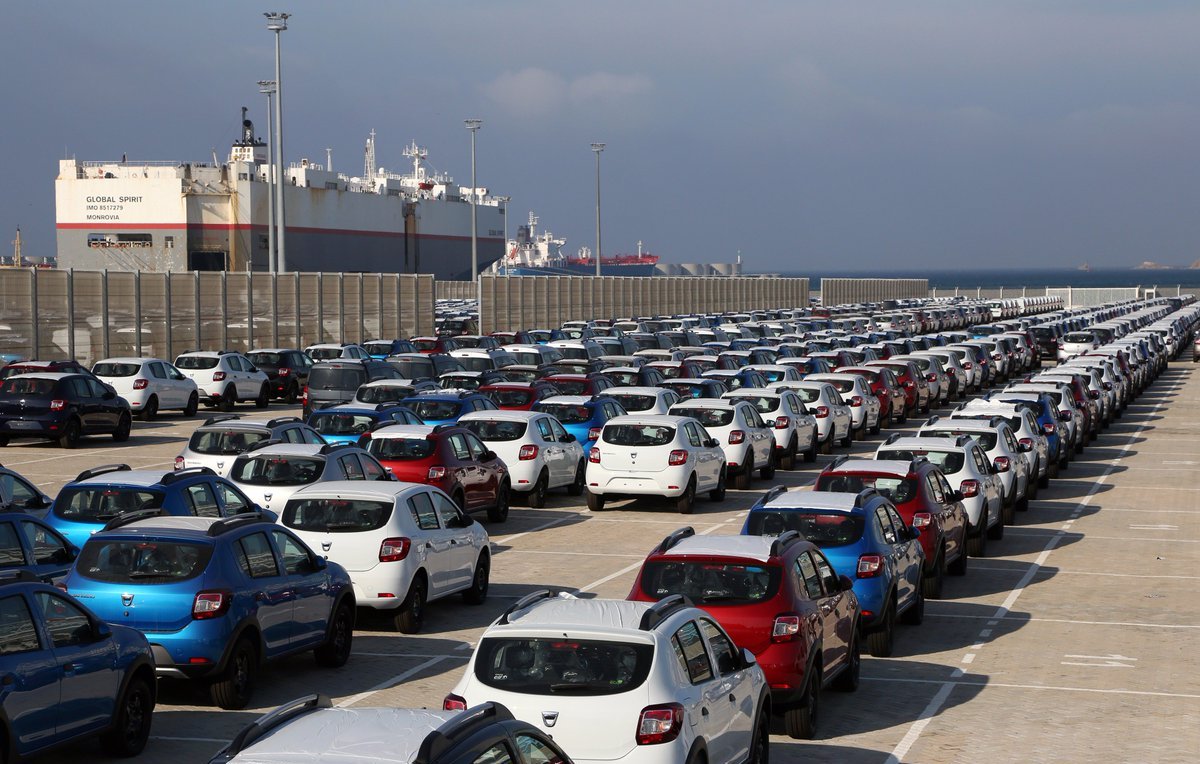 تظهر الصورة مشهداً لموقف سيارات كبير في ميناء، حيث تكون السيارات مصطفة بشكل منظم. يوجد مجموعة متنوعة من السيارات بألوان مختلفة، مثل الأبيض والأزرق والأحمر. في الخلفية، يمكن رؤية سفن راسية في الميناء تحت سماء زرقاء مع بعض السحب. يبدو أن المكان مخصص لتخزين السيارات، مما يوحي بأنه يعتمد على عمليات الشحن والنقل.