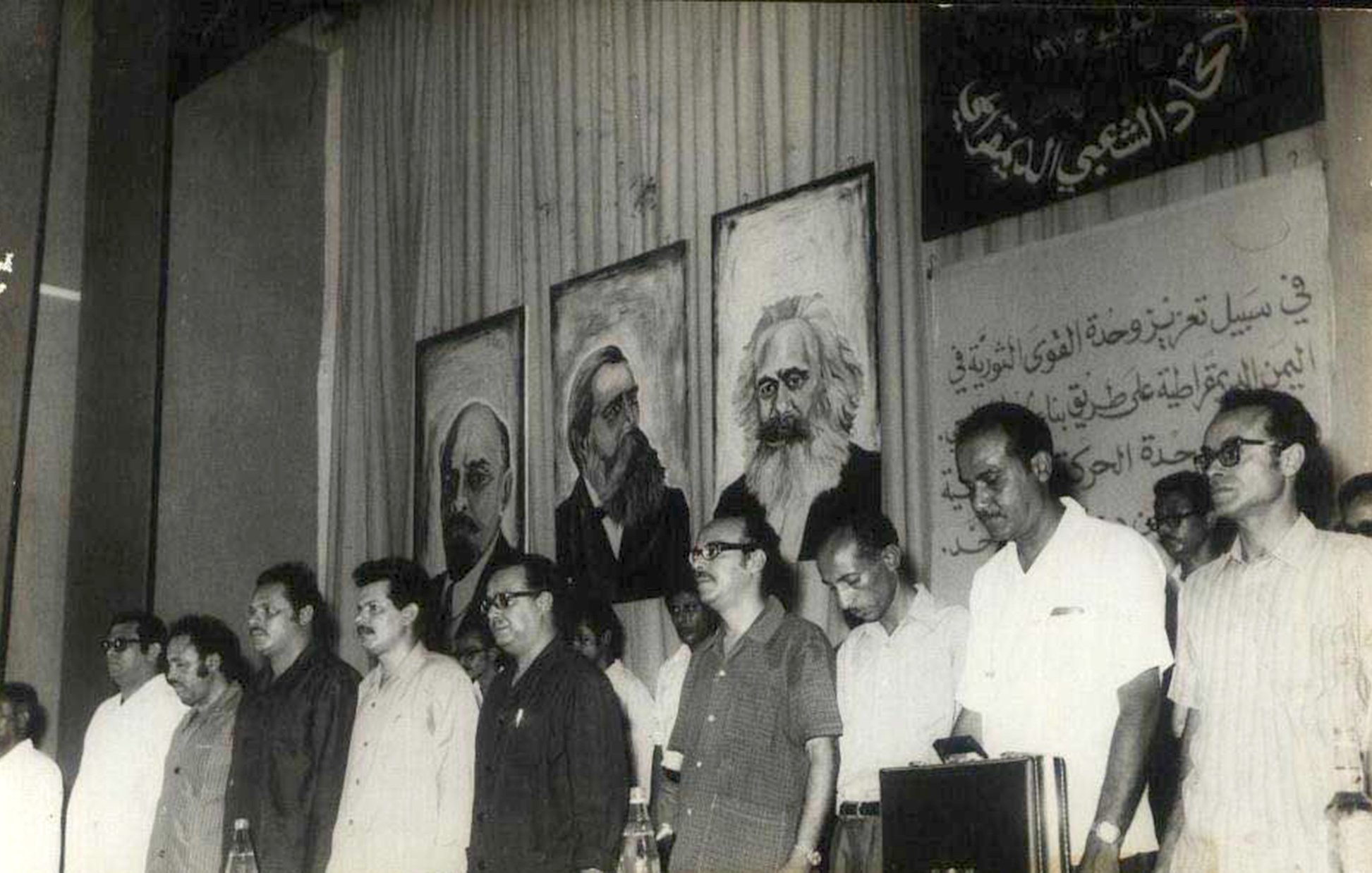L'image montre un groupe d'hommes se tenant debout en ligne, probablement lors d'un événement ou d'une conférence. Ils semblent pris dans un moment de réflexion ou de recueillement. À l'arrière-plan, on peut voir plusieurs portraits accrochés au mur, représentant des figures historiques ou intellectuelles, dont certaines sont bien connues. Le décor suggère un contexte politique ou idéologique, avec des inscriptions en arabe sur le mur qui renforcent ce thème. L'atmosphère générale semble sérieuse et engagée.