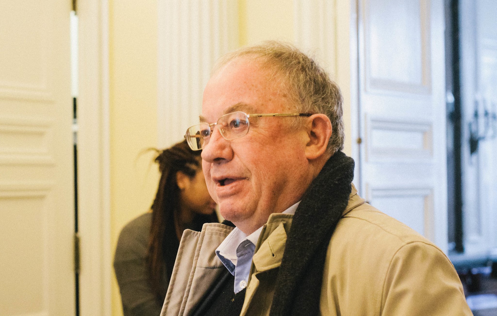 Homme âgé avec des lunettes, portant un manteau, dans un intérieur lumineux.