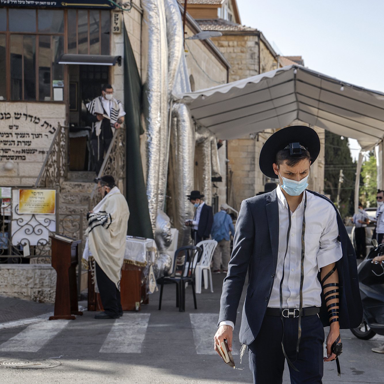 تظهر الصورة شابًا يرتدي بدلة مع قبعة، وهو يحمل هاتفًا محمولًا في يده. يتزين ذراعه بلفائف التوراة (تيفيلين)، ويبدو أنه في أحد الأماكن العامة. في الخلفية، توجد مجموعة من الأشخاص، وبعضهم يرتدي ملابس تقليدية، كما يظهر خيمة كبيرة على جانب الطريق. تتميز الصورة بأجواء حضرية مع وجود نباتات وبيئات سكنية.