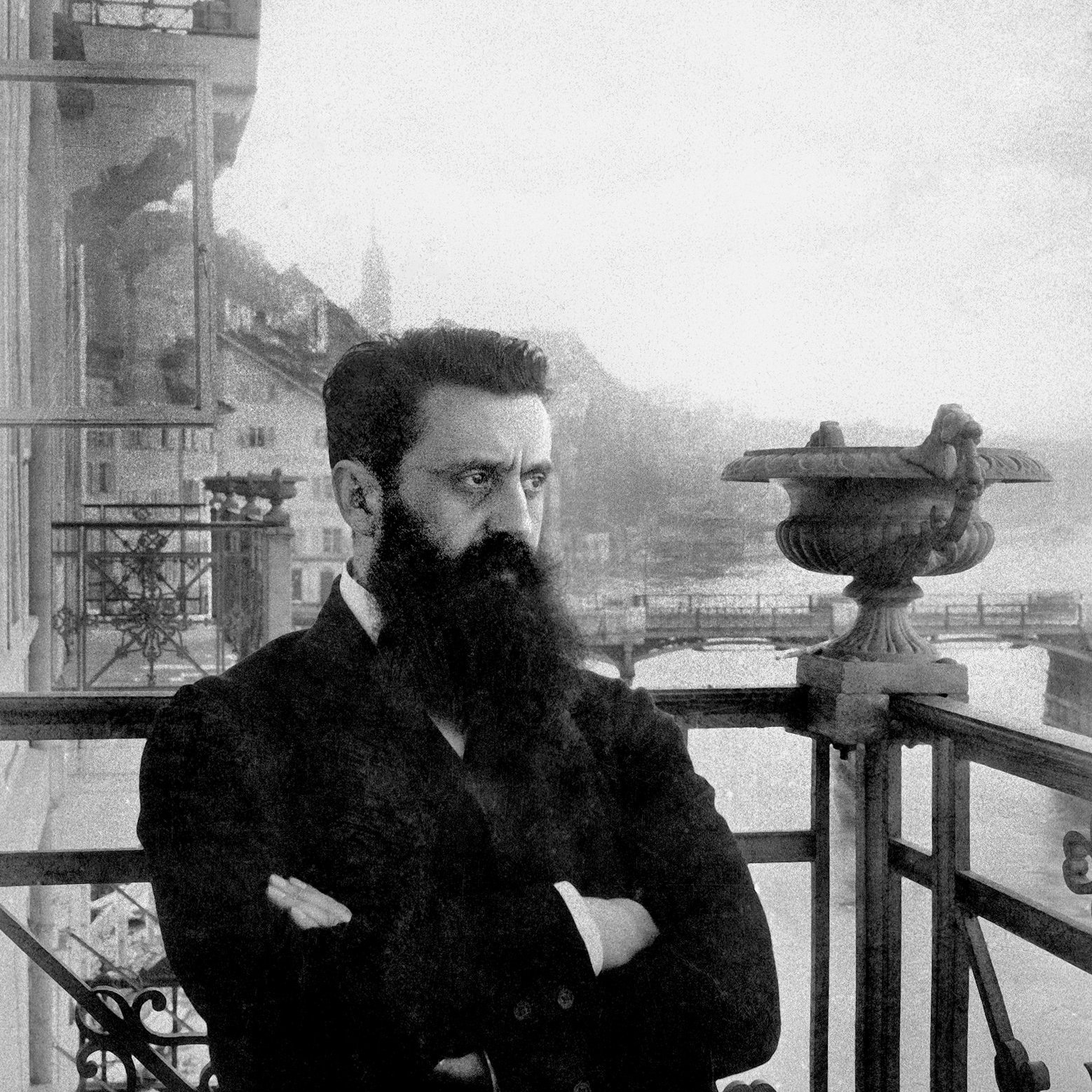 L'image montre un homme avec une barbe fournie, portant un costume sombre. Il est debout sur un balcon orné de balustrades métalliques, avec un regard pensif et sérieux. En arrière-plan, on aperçoit des montagnes et un plan d'eau, suggérant un paysage pittoresque. L'atmosphère est nostalgique, renforcée par le ton en noir et blanc de la photographie, ajoutant une dimension historique à la scène.