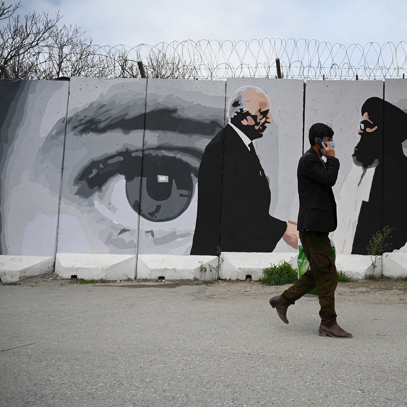 تظهر في الصورة حائطًا مزخرفًا برسوم جدارية لشخصيتين تتبادلان النظرات، حيث توجد عيون كبيرة في الخلفية تعكس مشاعر متباينة. في المقدمة، يمشي رجل يرتدي بدلة سوداء، ممسكًا بهاتفه المحمول. الحائط يتضمن أسلاك شائكة، مما يضيف إحساسًا بالانقسام أو التوتر. تبرز الألوان الرمادية والأبيض والأسود بشكل كبير، مما يضفي طابعًا دراماتيكيًا على المشهد.