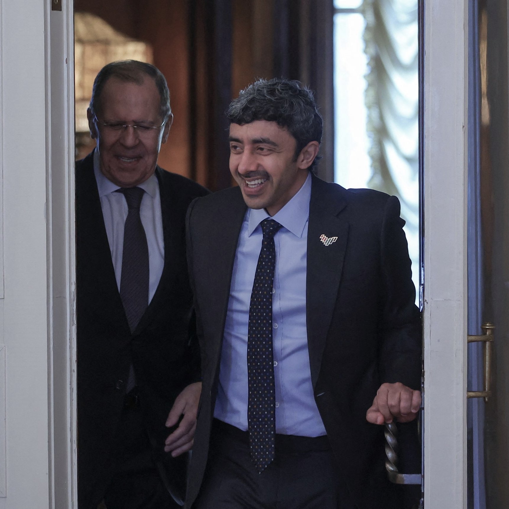 L'image montre deux hommes entrant dans une pièce. L'un d'eux sourit et semble de bonne humeur, tandis que l'autre a une expression plus neutre. Ils portent des costumes assortis et se trouvent dans un environnement élégant avec des éléments de décoration classique. La lumière naturelle entre par une fenêtre, ajoutant une ambiance chaleureuse à la scène.