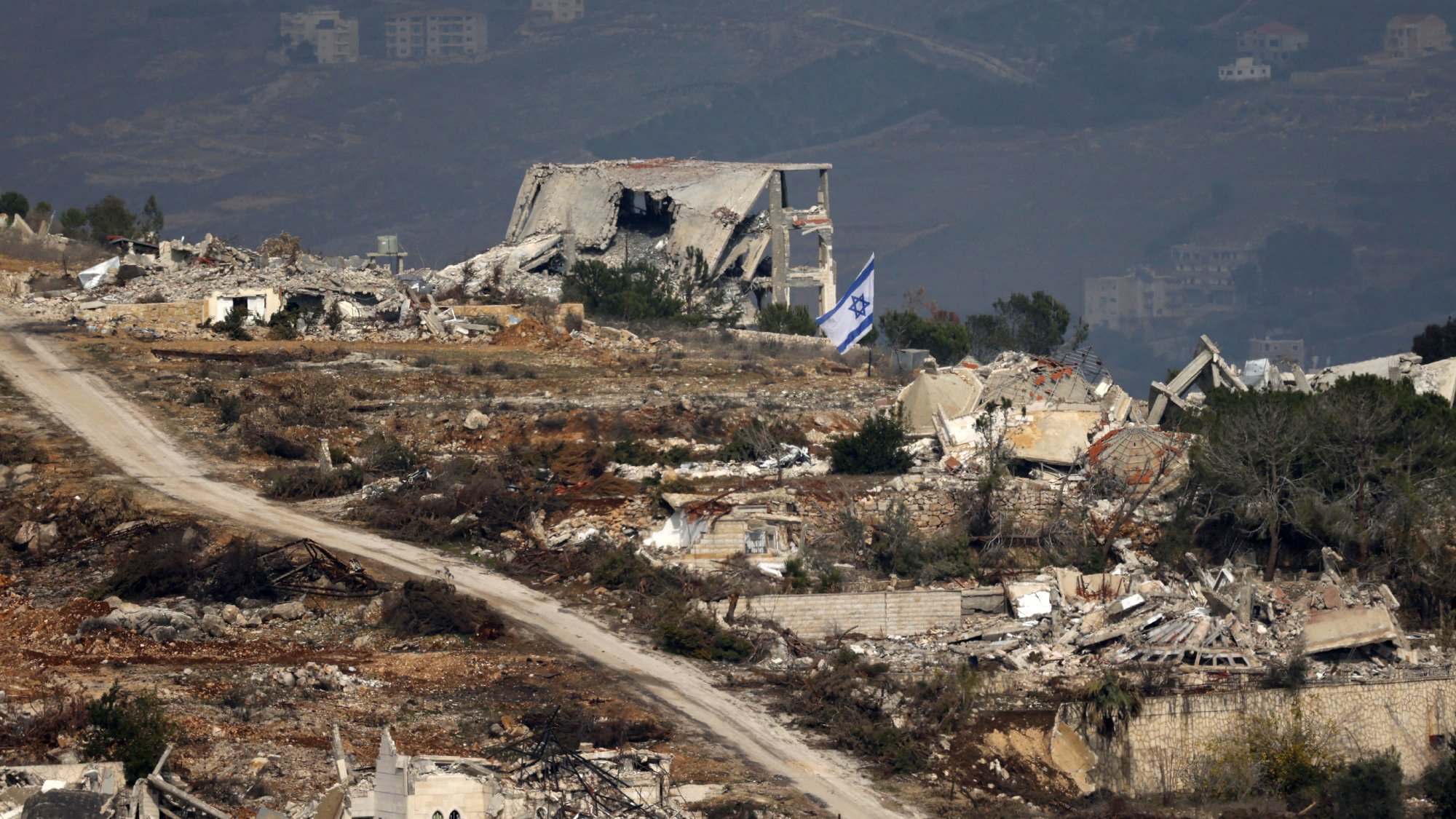 The image depicts a desolate landscape featuring extensive destruction, with several ruined buildings scattered throughout the area. In the foreground, there is a dirt road winding through the wreckage. Among the ruins, an Israeli flag can be seen prominently displayed, suggesting a connection to the location. The background shows additional structures and what appears to be a mountainous or hilly terrain, indicating a possibly rural setting. The overall scene conveys a sense of abandonment and turmoil.