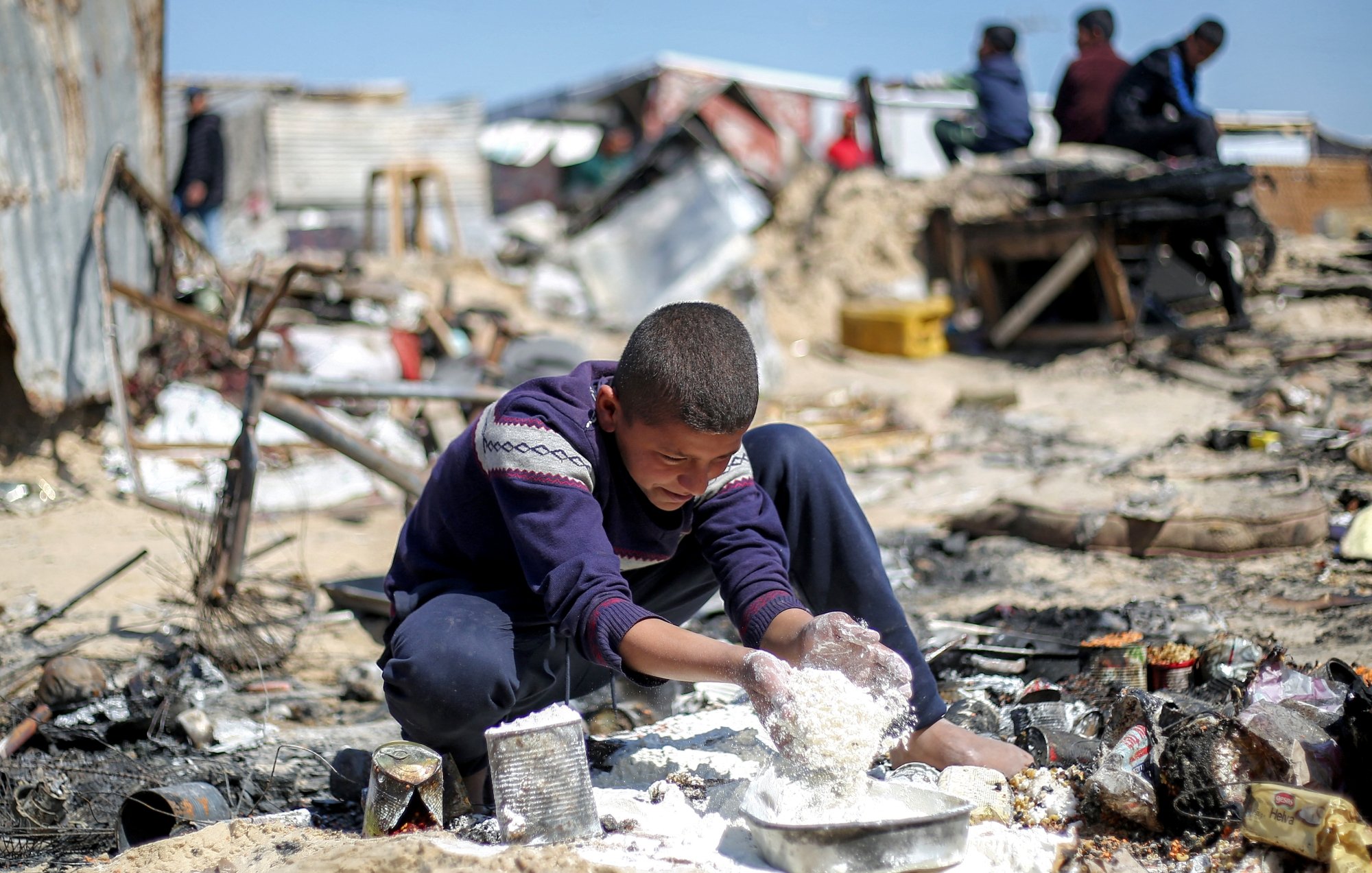 پسرکی در میان خرابه‌ها نشسته و مشغول بازی با مواد سفید رنگ است. فضای اطراف بسیار بی‌نظم و ویران است.
