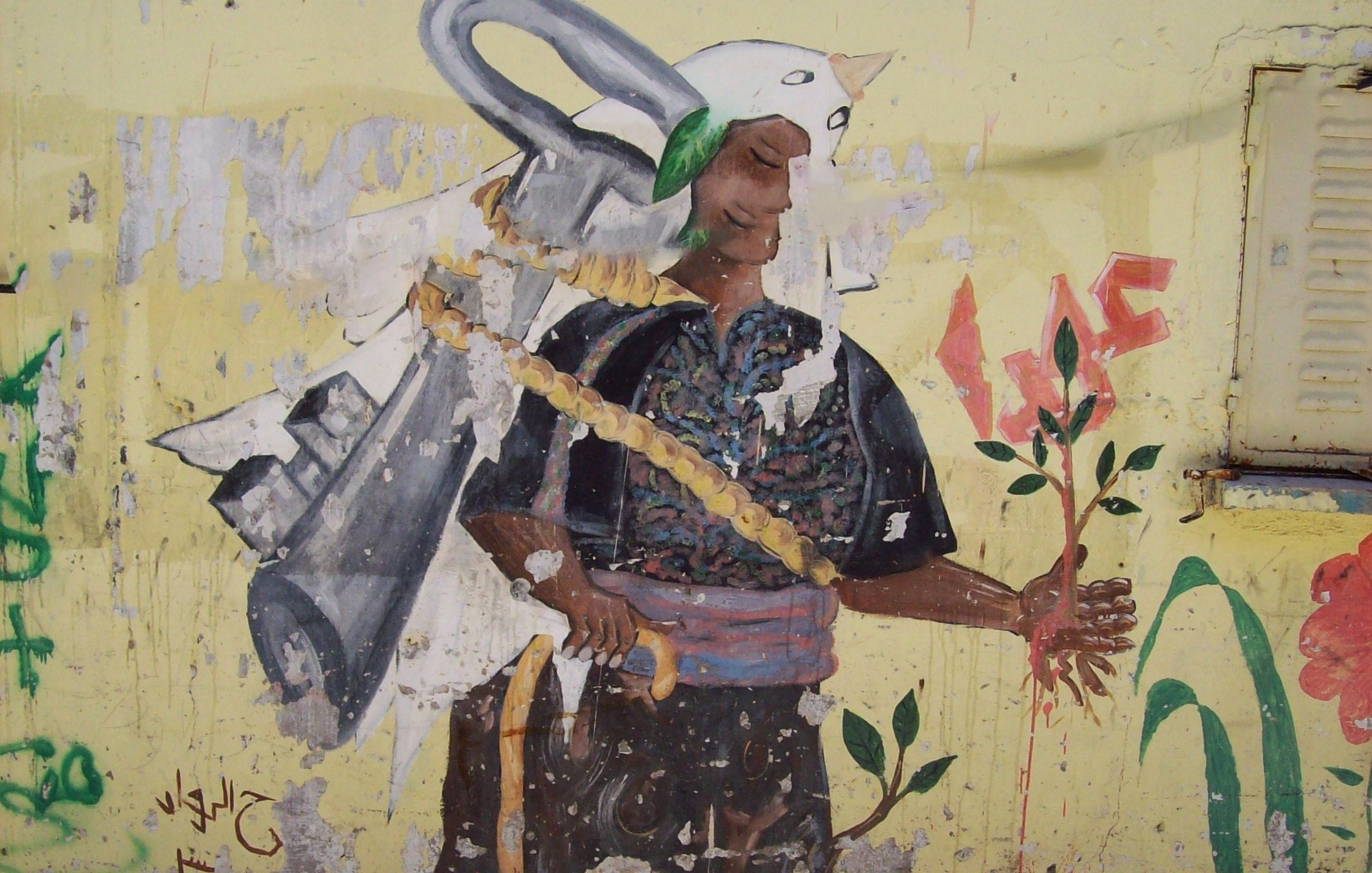 L'image montre une fresque sur un mur. On y voit un personnage portant un costume traditionnel, avec un air déterminé. Ce personnage tient une plante dans une main, symbolisant la vie et la nature. Sur son dos, on peut apercevoir une sorte d'outil ou d'équipement qui pourrait symboliser le travail ou un engagement envers la terre. Le fond est de couleur jaune, ce qui attire l'œil et met en avant le contraste avec les couleurs du personnage. Des inscriptions et des graffitis en arrière-plan ajoutent du caractère à l'œuvre.