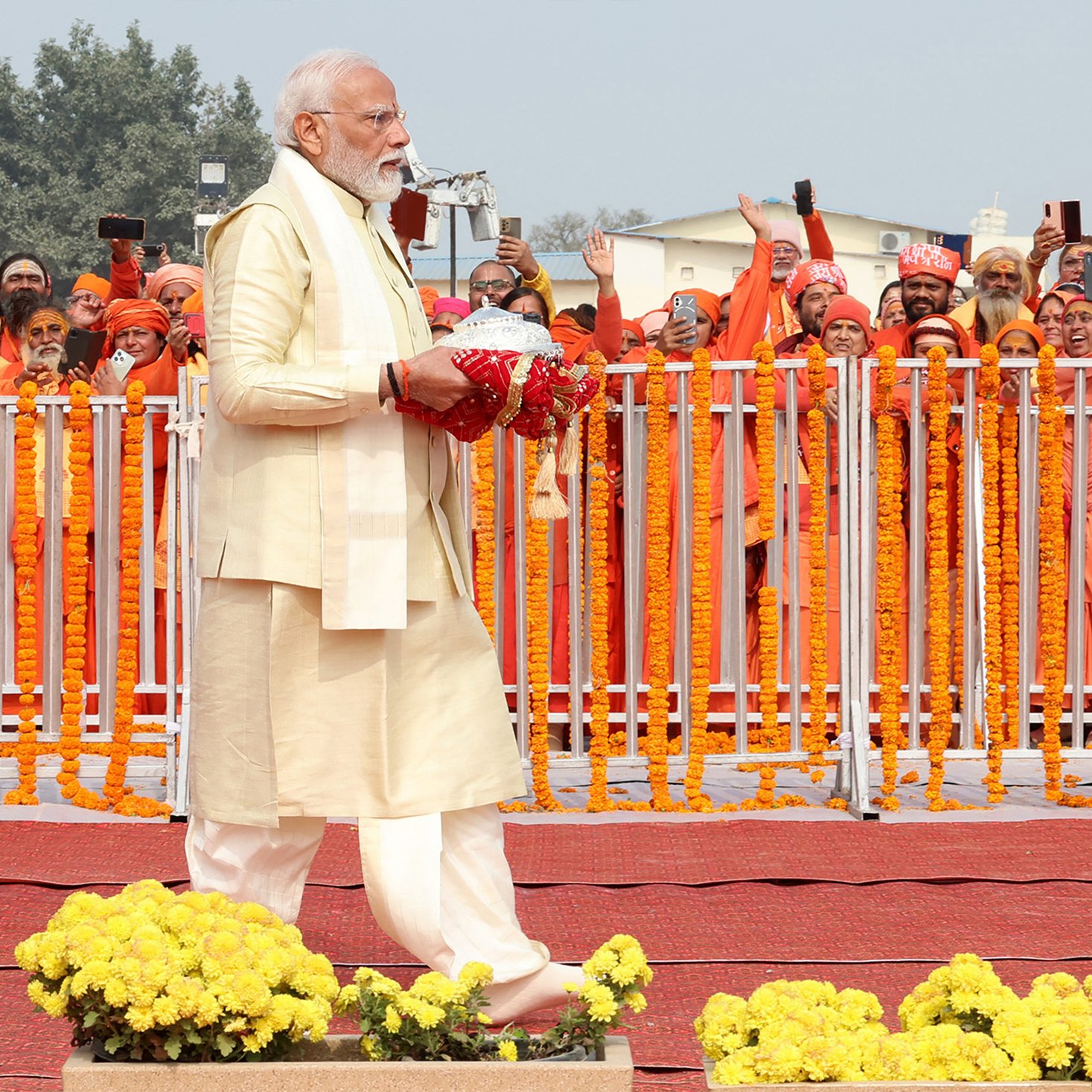 L'image montre une scène publique, probablement lors d'un événement officiel. Un homme portant une tenue traditionnelle claire, avec une longue barbe et des cheveux gris, marche sur une estrade. Il tient un objet dans ses mains, peut-être un bouquet ou une offrande. En arrière-plan, un grand groupe de personnes vêtues d'habits oranges l'acclament, certaines tenant des téléphones pour prendre des photos. L'environnement est décoré de fleurs et de guirlandes. L'atmosphère semble festive et respectueuse.
