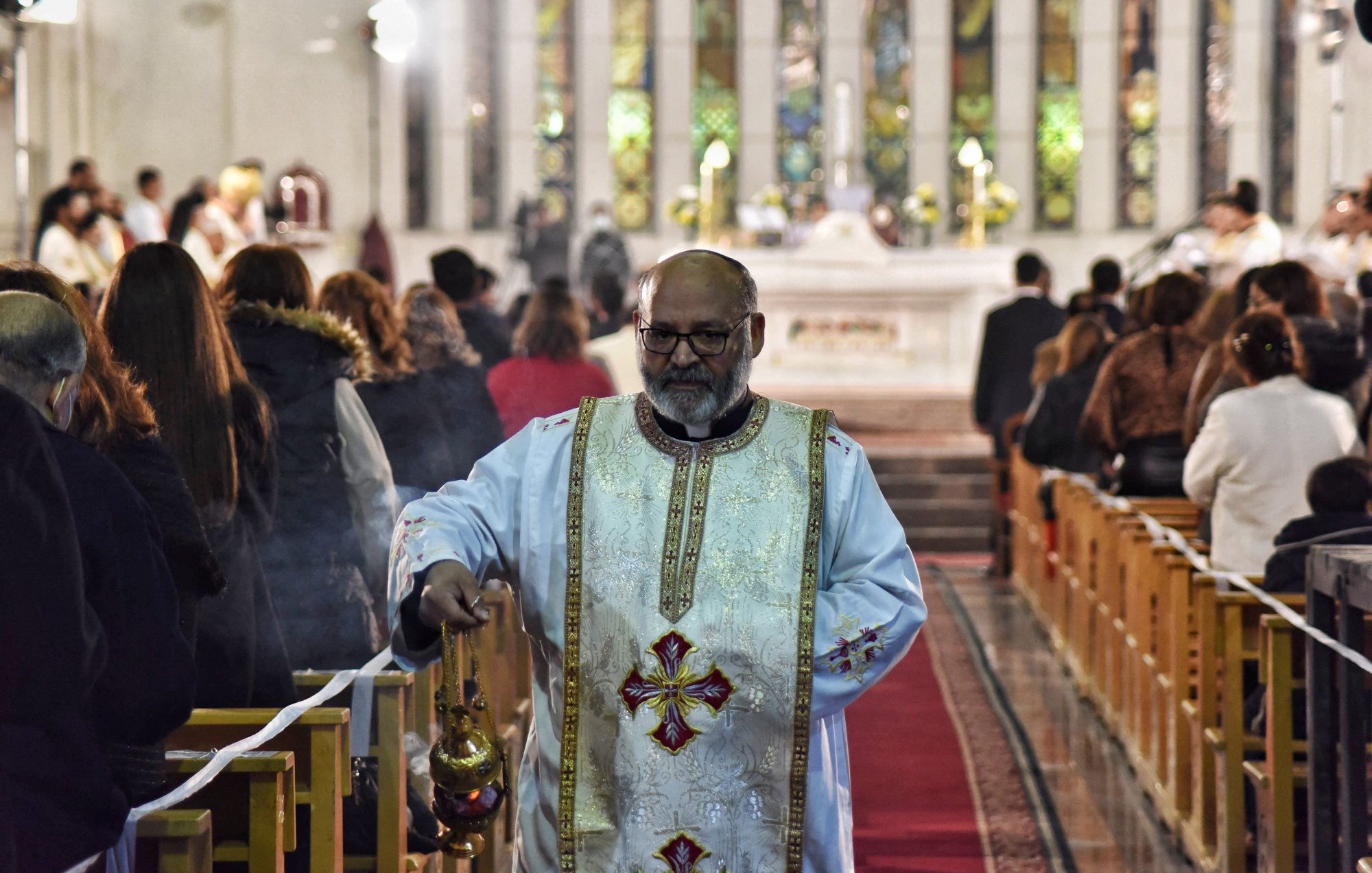 Un prêtre en tenue liturgique, portant un encensoir, dans une église pleine de fidèles.