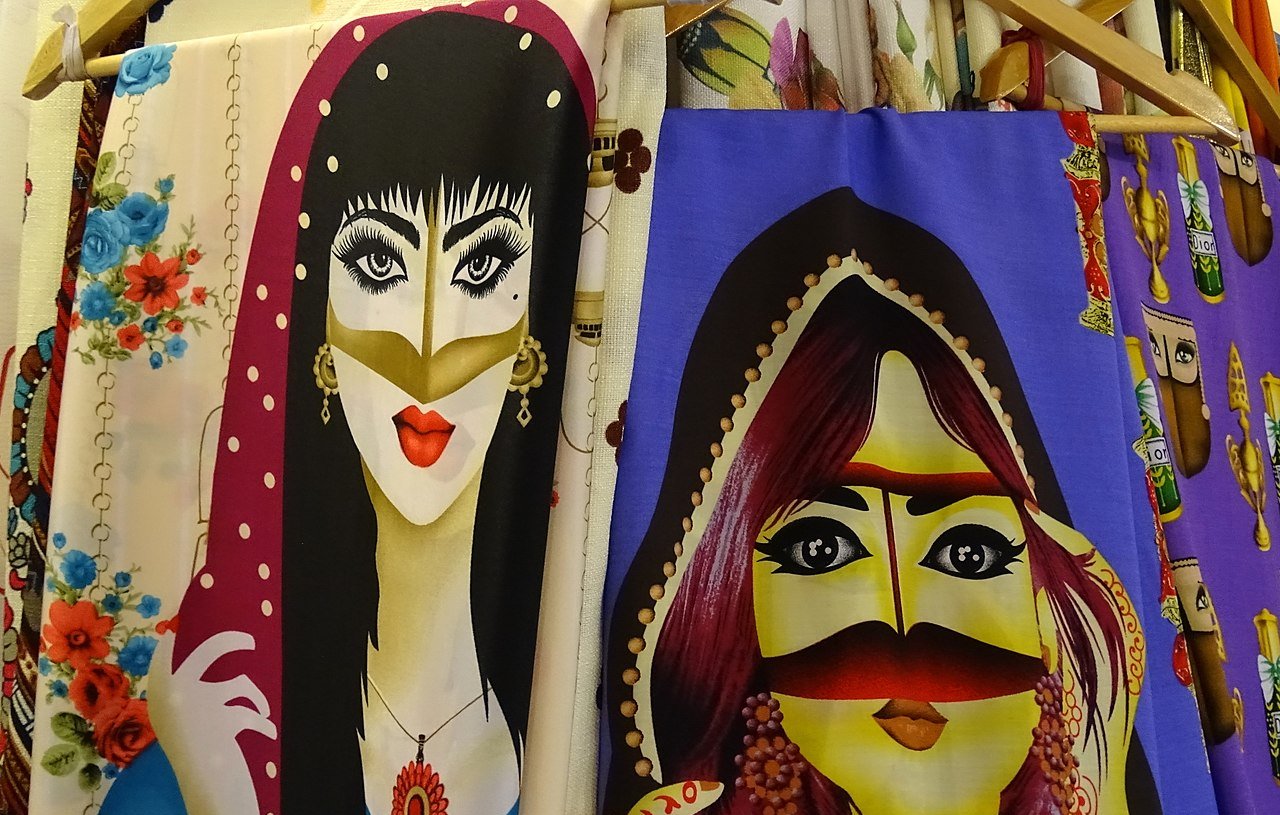 تظهر في الصورة رسومات نسائية ملونة على قماش. الشخصيات المحتواة في الرسومات ترتدي أزياء تقليدية وتتميز بتفاصيل دقيقة في الوجه والعينين. الألوان زاهية، حيث تم استخدام درجات متنوعة من الألوان لجذب الانتباه، مما يعكس ثقافة معينة أو نمط فني مميز. يمكن رؤية بعض الزينة والإكسسوارات حول الشخصيات، مما يضيف لمسة من الأناقة.