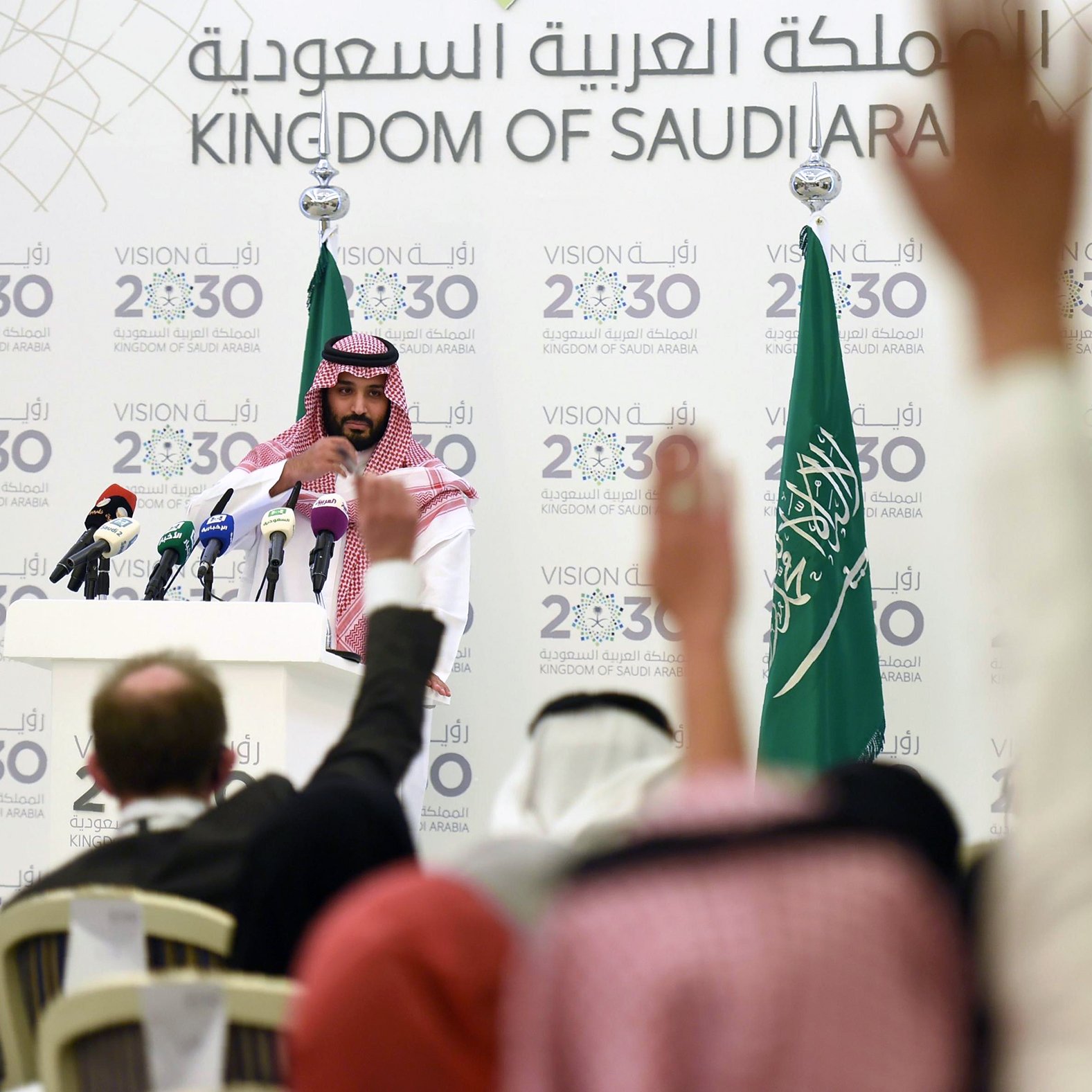 در این تصویر، یک مرد جوان با لباس سنتی سعودی و عمامه، در حال سخنرانی در مقابل میکروفن‌ها و دوربین‌های خبری است. او در پس‌زمینه‌ای شامل پرچم‌های عربستان سعودی و شعارهایی درباره "چشم‌انداز ۲۰۳۰" قرار دارد. تعدادی از افراد نشسته در سالن، دست‌های خود را برای پرسش بالا برده‌اند. فضا به وضوح نشان‌دهنده یک کنفرانس خبری یا رویداد رسمی است.