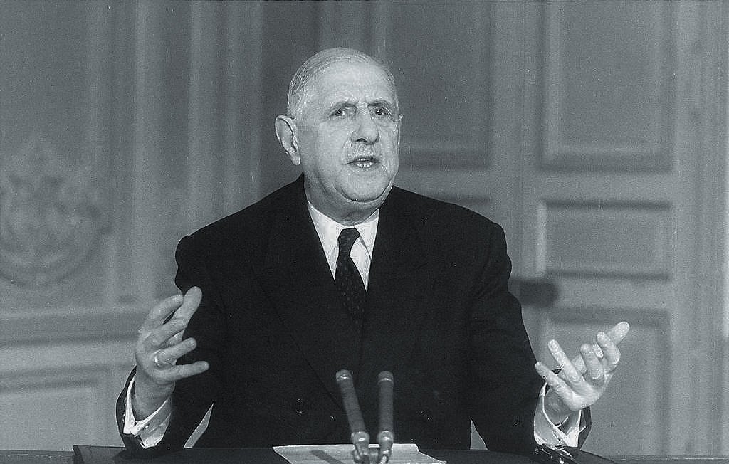The image depicts a man in a formal suit seated at a desk, speaking with his hands raised expressively. He appears to be delivering a speech or addressing an audience. The setting suggests a formal environment, likely a governmental or political space, with a plain background that emphasizes the speaker. The monochromatic tone gives the image a historical feel, suggesting it may be from a past era.
