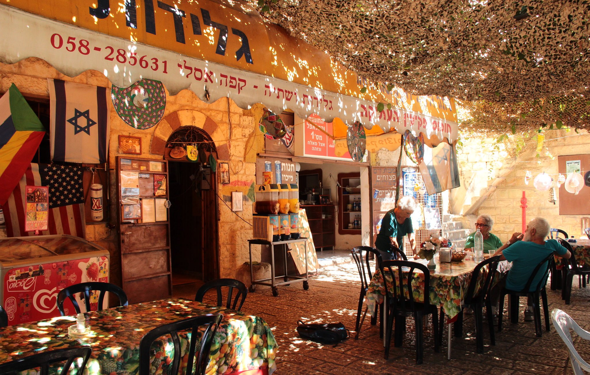 L'image montre un café ou une taverne en plein air, avec des tables et des chaises noires disposées sur un sol en pierre. Le lieu semble très accueillant, avec des nappes colorées sur les tables. Au-dessus, un toit en toile de camouflage laisse passer un peu de lumière. On peut voir des drapeaux accrochés aux murs, probablement des symboles de différentes nationalités. Des personnes assises discutent tranquillement dans cet environnement convivial. L'architecture est typique des bâtiments anciens, ajoutant une touche authentique à l'ambiance.
