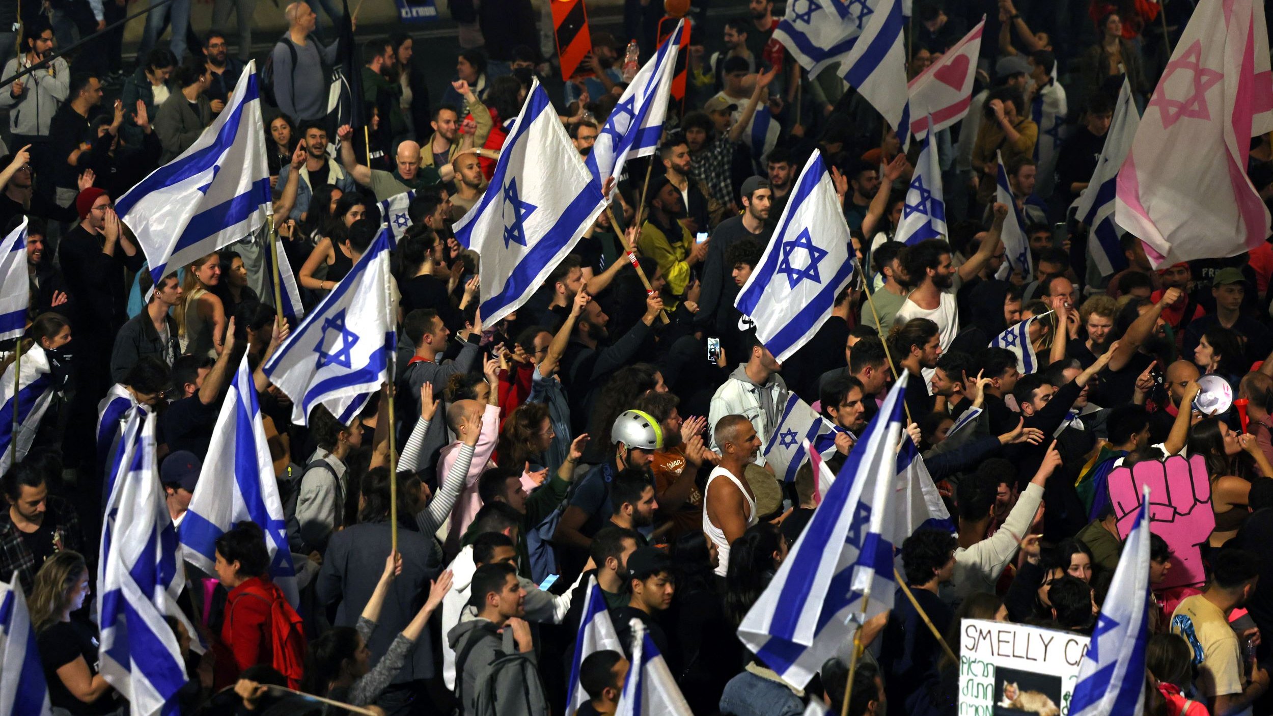 La imagen muestra una multitud de personas en una manifestación, sosteniendo banderas de Israel. Hay un ambiente de entusiasmo y participación, con la gente ondeando las banderas en un espacio al aire libre. Algunas personas parecen estar animadas, mientras que otras sostienen carteles. El contexto sugiere que podría tratarse de una protesta o un evento significativo relacionado con Israel.