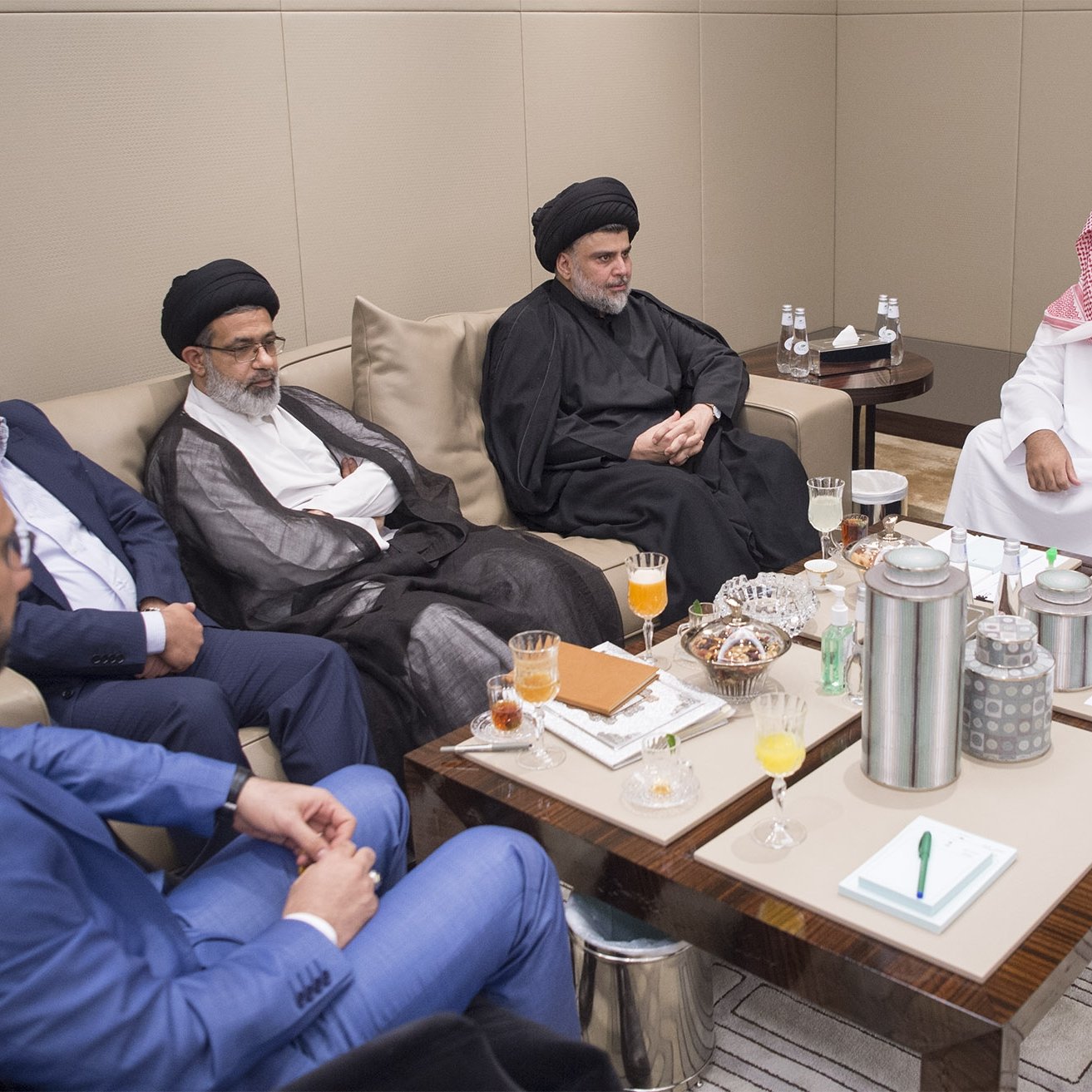 L'image montre un groupe de personnes assises autour d'une table dans une pièce moderne et élégante. Ils semblent engagés dans une discussion sérieuse. Sur la table, il y a des boissons et des collations. Les participants portent des vêtements formels, certains avec des vêtements traditionnels, ce qui souligne le contexte culturel de la réunion. L'ambiance est professionnelle et conviviale.