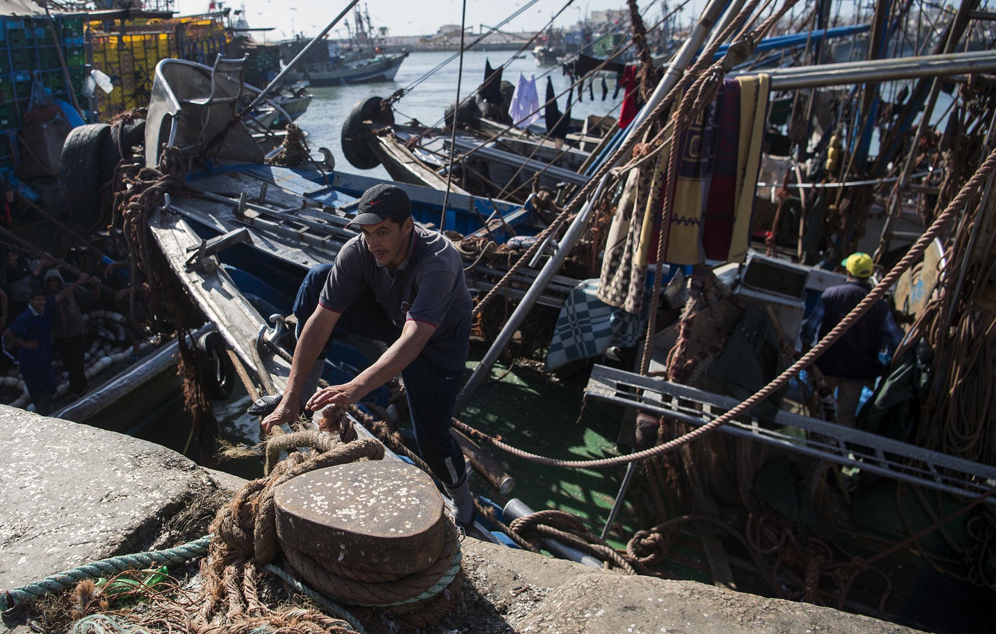 L'image montre un port de pêche animé, avec des bateaux de pêche amarrés. Un homme, vêtu d'un t-shirt et d'une casquette, est visible en train de travailler sur le quai, s'occupant de cordages. En arrière-plan, on peut voir d'autres bateaux et une atmosphère maritime. Le paysage évoque une activité portuaire typique, avec plusieurs éléments liés à la pêche.