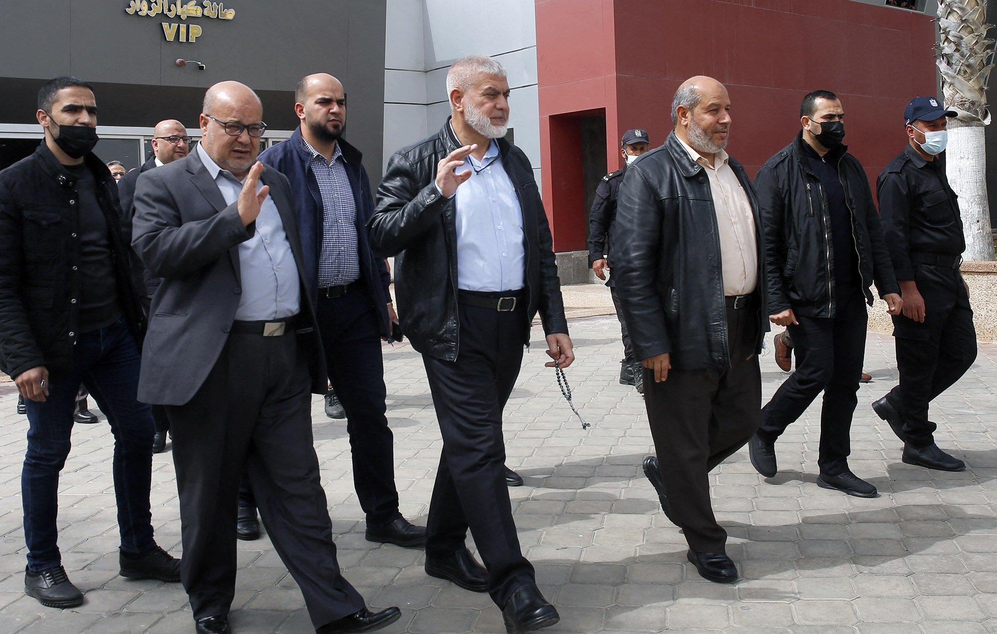 Nell'immagine si vede un gruppo di uomini che camminano insieme all'aperto. Alcuni di loro indossano giacche di pelle e altri abiti formali. Alcuni uomini indossano mascherine, mentre altri no. Sembrano impegnati in una conversazione e uno di loro alza la mano in un gesto di saluto. Sullo sfondo si intravedono strutture architettoniche moderne e una pavimentazione in pietra. L'atmosfera sembra formale e organizzata.