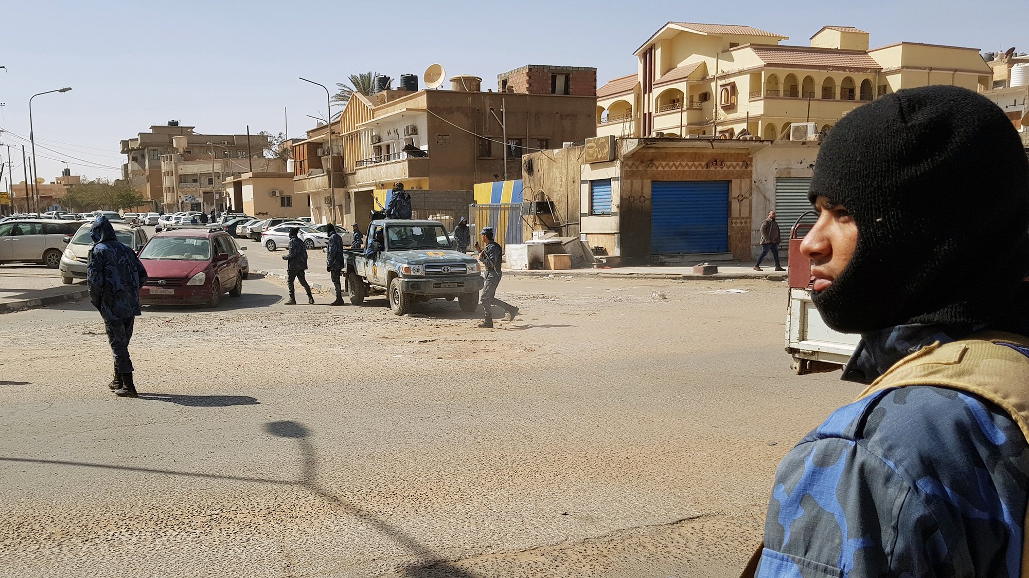 L'image montre une scène urbaine où des policiers armés sont présents dans une rue. On peut voir plusieurs membres des forces de l'ordre, certains portant des uniformes camouflés et des masques, tout en étant attentifs à leur environnement. Il y a des véhicules de police, ainsi que des voitures stationnées à proximité. En arrière-plan, on aperçoit des bâtiments de style architectural varié. L'atmosphère semble tendue, évoquant un contexte de sécurité renforcée.