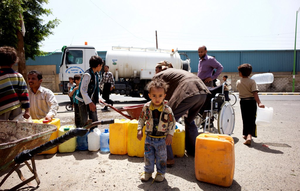 L'image montre une scène animée dans une zone urbaine, où plusieurs personnes sont rassemblées autour d'un camion-citerne. Des adultes et des enfants transportent des récipients pour collecter de l'eau. On remarque un jeune enfant au premier plan, portant une veste colorée, se tenant debout parmi d'autres personnes qui s'affairent. En arrière-plan, on aperçoit un homme en fauteuil roulant qui semble aider. Les couleurs des récipients sont vives, principalement jaunes et bleus, et l'atmosphère illustre un besoin urgent d'approvisionnement en eau.