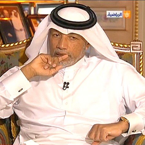 L'image montre un homme assis sur un fauteuil, habillé en costume traditionnel arabe, avec un headdress. Il a une expression pensive et semble concentré, en train de penser ou de réfléchir. En arrière-plan, on peut voir des photographies accrochées au mur, ajoutant une touche personnelle à l'environnement. L'atmosphère semble être celle d'une interview ou d'une discussion sérieuse.