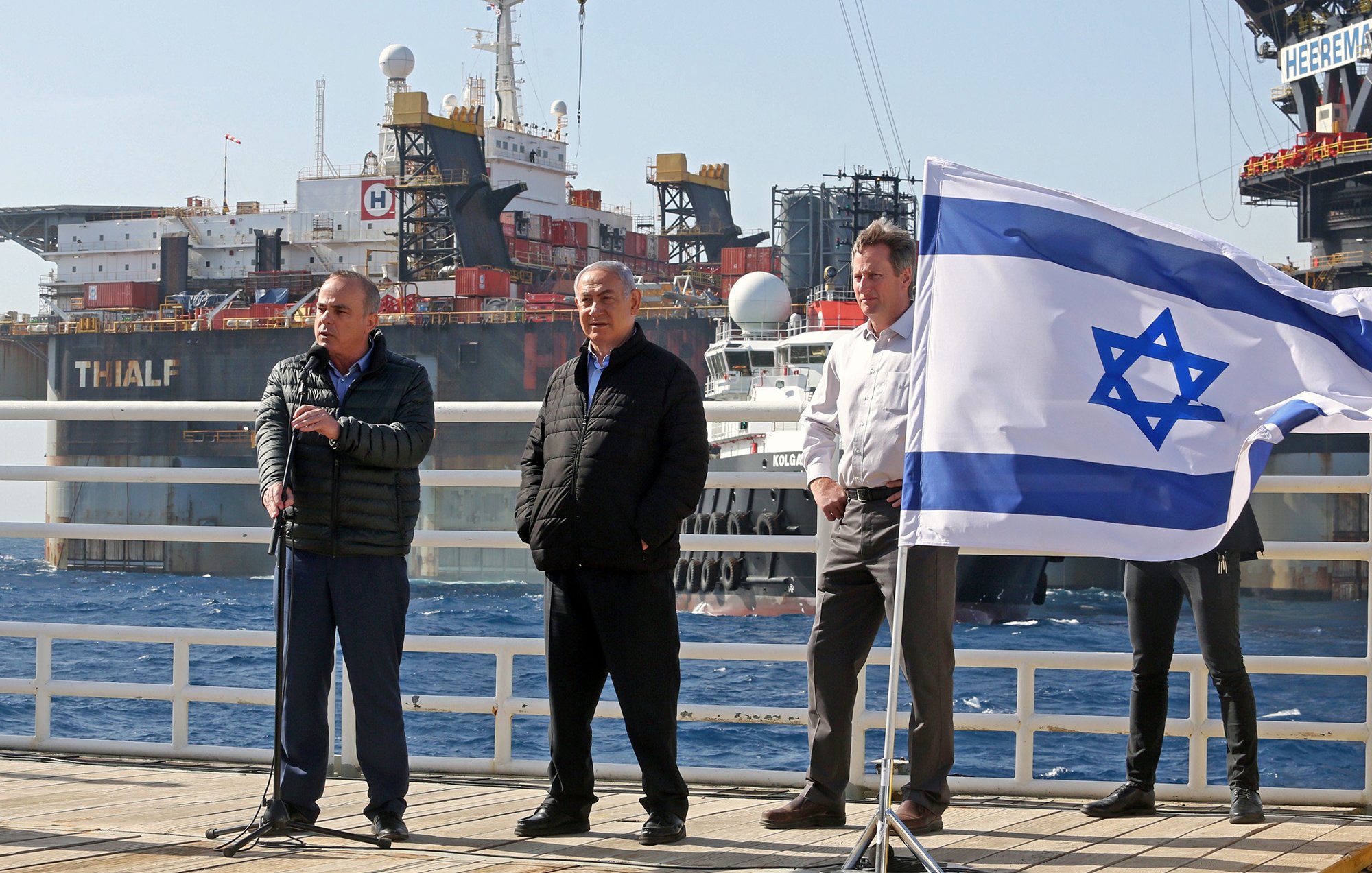 L'image montre plusieurs personnes sur un quai, en face de navires en arrière-plan. À gauche, un homme s'exprime avec un micro, tandis qu'un autre homme se tient à ses côtés, visiblement en position d'écoute. Un troisième homme est également présent. Au premier plan, un drapeau israélien flotte, ajoutant une touche patriotique à la scène. Les navires derrière eux semblent être engagés dans des opérations maritimes, suggérant un contexte lié à l'industrie maritime ou énergétique. L'ambiance paraît formelle, indiquant probablement un événement officiel ou une annonce importante.