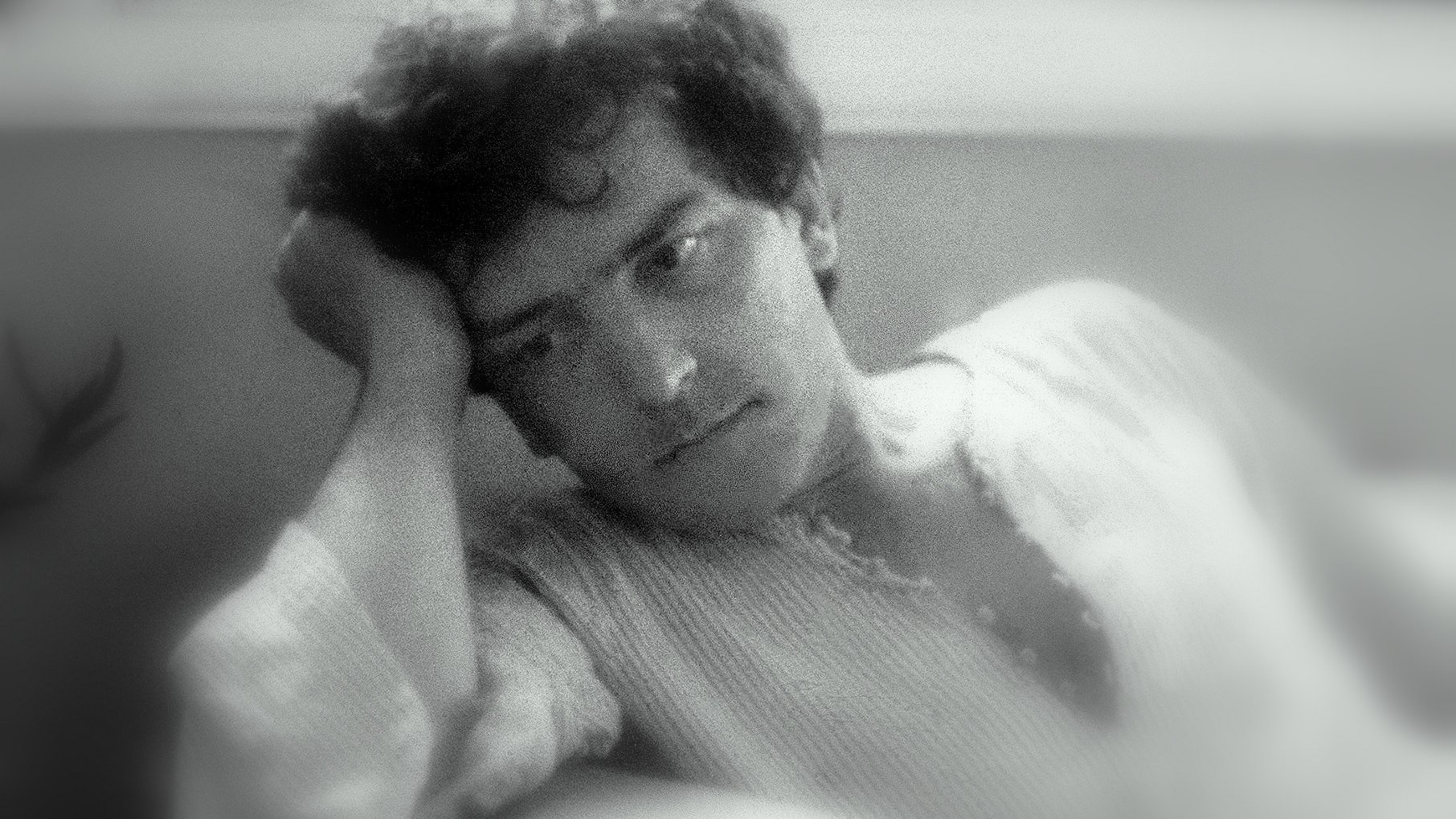 L'image montre un homme pensif, assis dans un endroit confortable. Il a les cheveux bouclés et un air réfléchissant, avec une main soutenant sa tête. Son expression semble mélancolique ou pensive. L'éclairage doux donne une ambiance nostalgique et calme à la scène. L'arrière-plan flou contribue à centrer l'attention sur son visage.