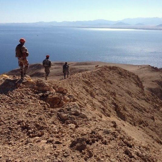 L'image montre un paysage désertique avec des militaires en patrouille sur une crête surplombant une étendue d'eau. Le terrain est aride et rocheux, avec des montagnes en arrière-plan. Les soldats portent des uniformes et des casques, et semblent observer l'horizon, avec un ciel clair et ensoleillé qui ajoute à l'atmosphère de la scène.