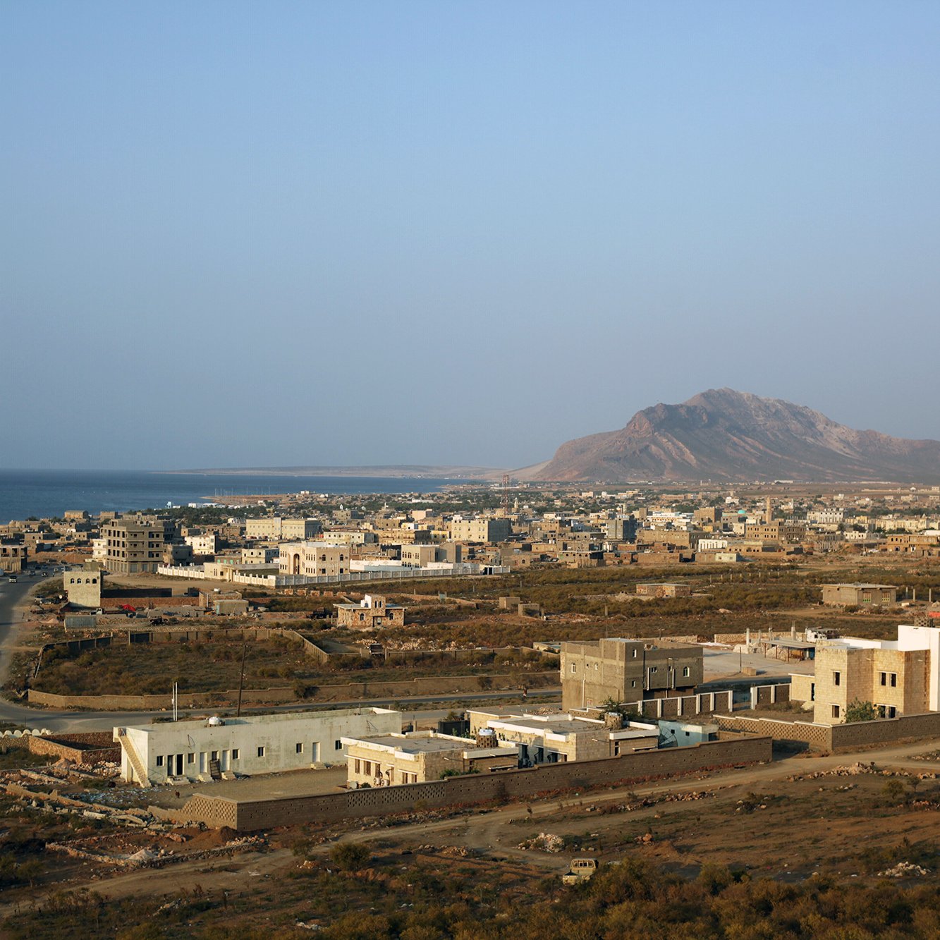 The image depicts a coastal landscape with a view of a small town or village near the ocean. In the foreground, there are a few buildings, some of which are modern, while others appear more traditional. The road winds through the landscape, leading towards the sea. In the background, a mountain rises against a clear sky, adding to the natural beauty of the scene. The overall atmosphere suggests a quiet, possibly rural environment with a mix of natural and man-made elements.