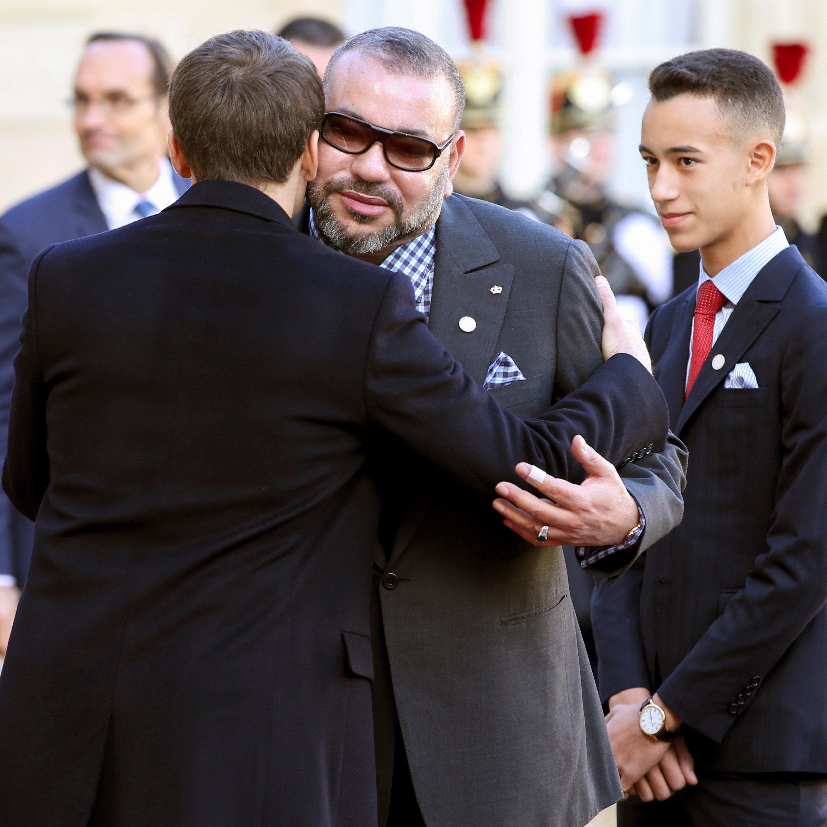 L'image montre une rencontre officielle entre deux hommes. L'un des hommes, vêtu d'un costume élégant, semble accueillir l'autre avec une accolade amicale. L'autre homme, portant des lunettes de soleil et un costume gris, se tient à côté d'un jeune garçon en costume sombre et cravate. En arrière-plan, on aperçoit des gardes en uniforme, ce qui indique un contexte cérémonial. L'atmosphère semble conviviale et respectueuse.