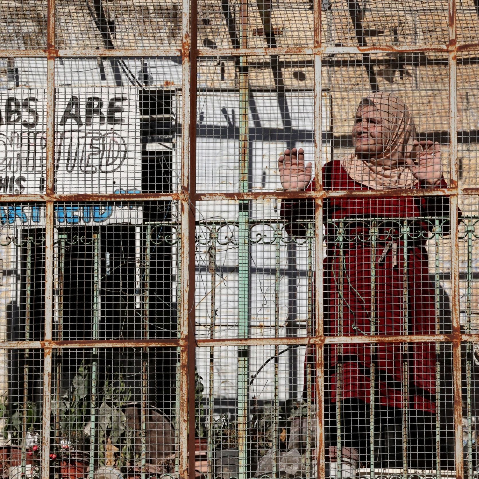 L'image montre une femme se tenant derrière une structure en métal grillagée. Elle porte un habit traditionnel et semble regarder à travers les barreaux. En arrière-plan, on aperçoit un panneau avec des mots en anglais qui semblent exprimer un message, mais son contenu exact reste obscurci par la grille. La scène évoque des thèmes de confinement et de séparation, avec une atmosphère de tension ou de défi face à une réalité difficile. Des plantes sont visibles dans l'environnement.
