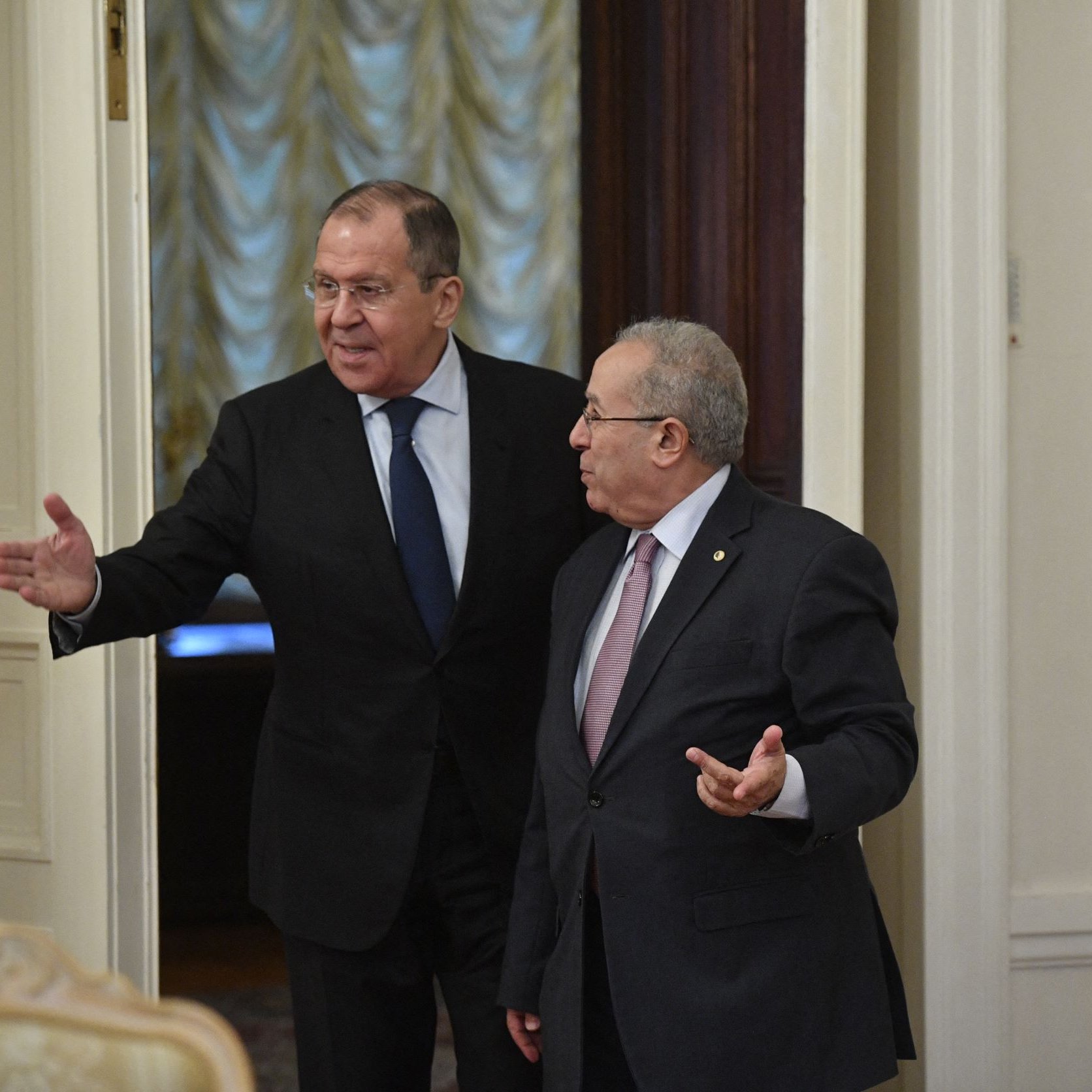 L'image montre deux hommes en train de discuter dans une pièce élégante. L'un d'eux, à gauche, porte un costume sombre et une cravate, et semble parler avec gestuelle. L'autre homme, à droite, est en costume clair et écoute attentivement. La pièce est bien décorée, avec des murs aux teintes claires et des rideaux d'apparence luxueuse.