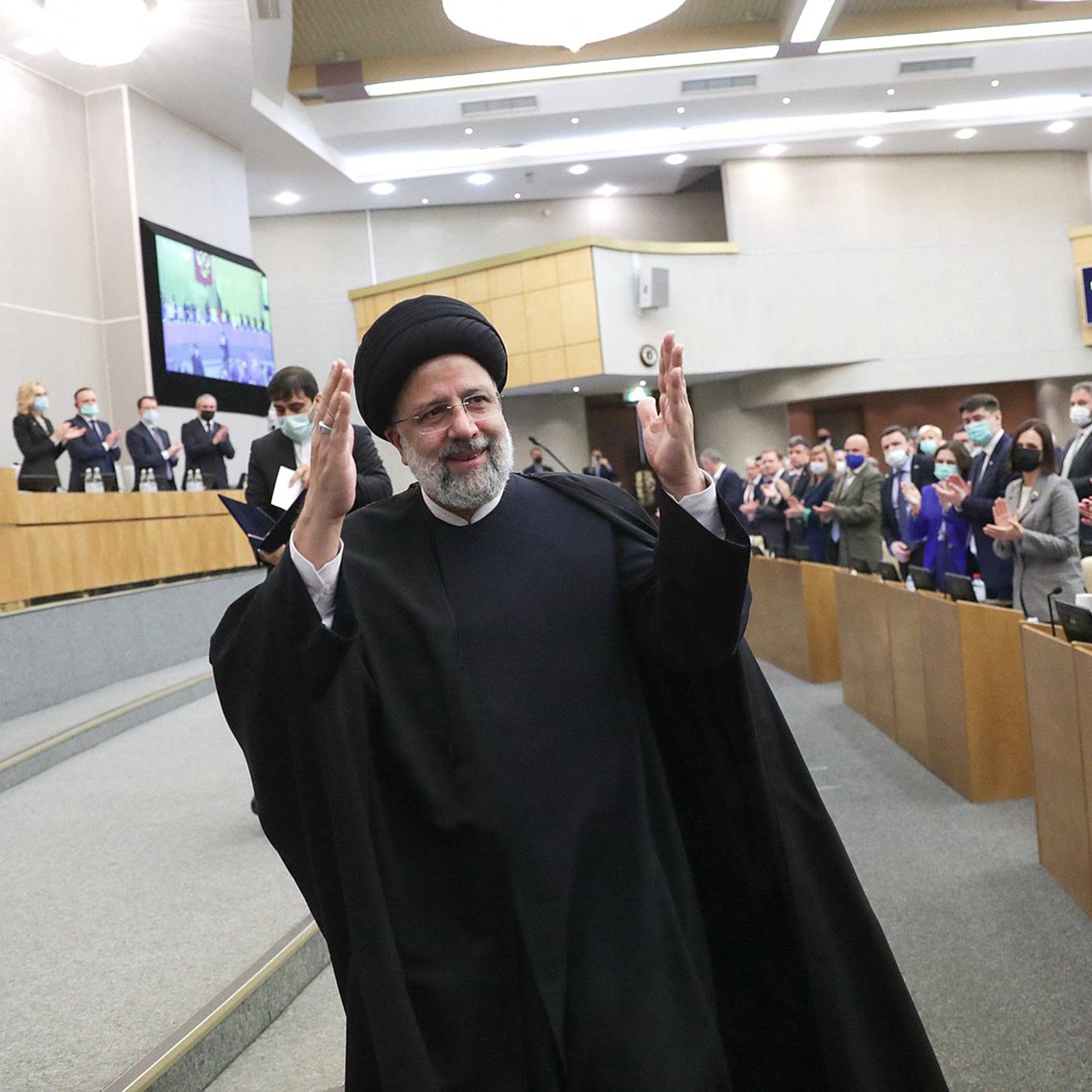 L'image montre un homme en habit traditionnel, portant un turban noir, qui semble être en train de saluer un public. Il se tient au centre d'une salle où plusieurs personnes applaudissent. L'environnement est formel, typique d'un événement politique ou d'une réunion officielle, avec des sièges et des écrans visibles en arrière-plan. Les participants portent également des masques de protection, indiquant une attention à la santé publique.
