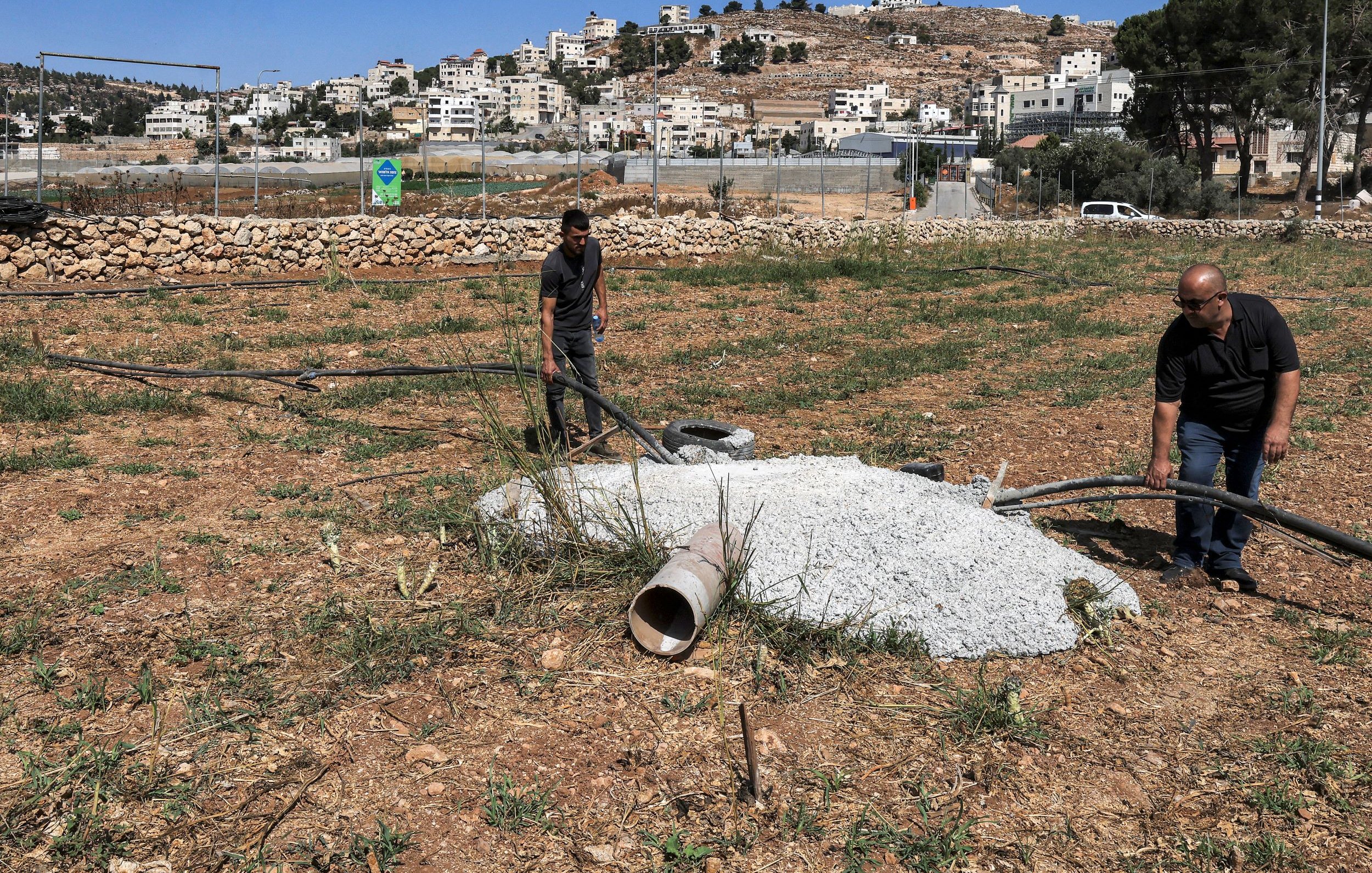 L'image montre deux hommes travaillant sur un terrain en plein air, probablement dans une zone rurale. Ils manipulent des tuyaux en plastique et se trouvent à proximité d'un tas de ciment ou de béton, ainsi qu'un petit tuyau qui sort du sol. À l'arrière-plan, on peut apercevoir des bâtiments et des collines. La végétation est clairsemée, et le sol semble fraîchement labouré ou non entretenu.