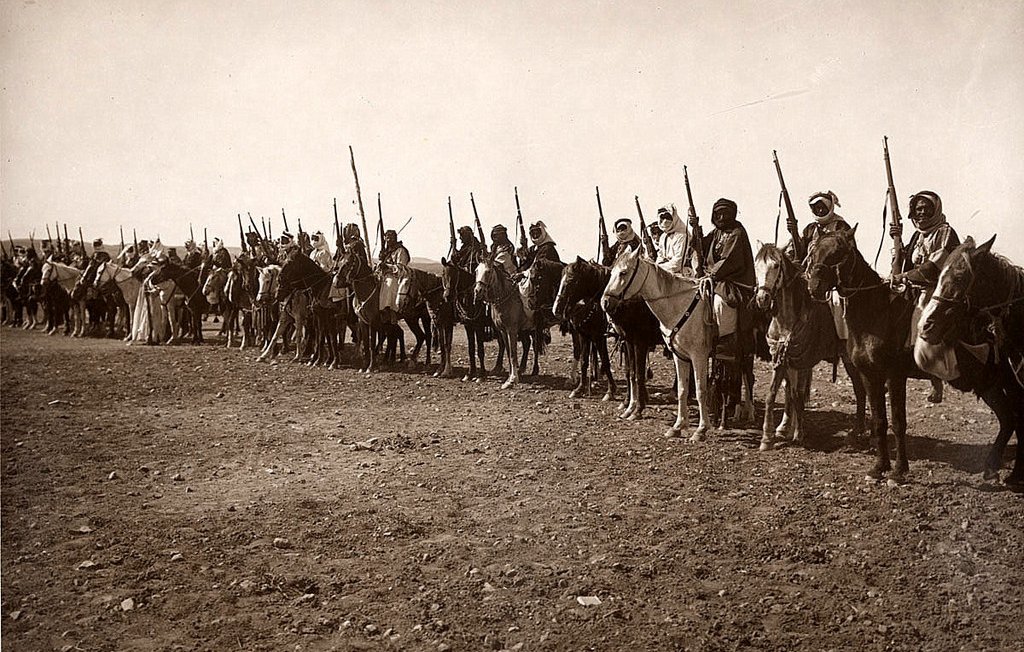 L'image montre une longue ligne de cavaliers en rang, tous montés sur des chevaux. Chaque cavalier est équipé d'une lance ou d'un bâton, et beaucoup portent des vêtements traditionnels. L'arrière-plan semble désertique, avec un sol rugueux et peu de végétation. L'atmosphère de l'image évoque une scène historique ou militaire, mettant en avant des guerriers ou des membres d'une tribu. Les cavaliers adoptent une posture fière et attentive, prêts pour une action ou une procession.